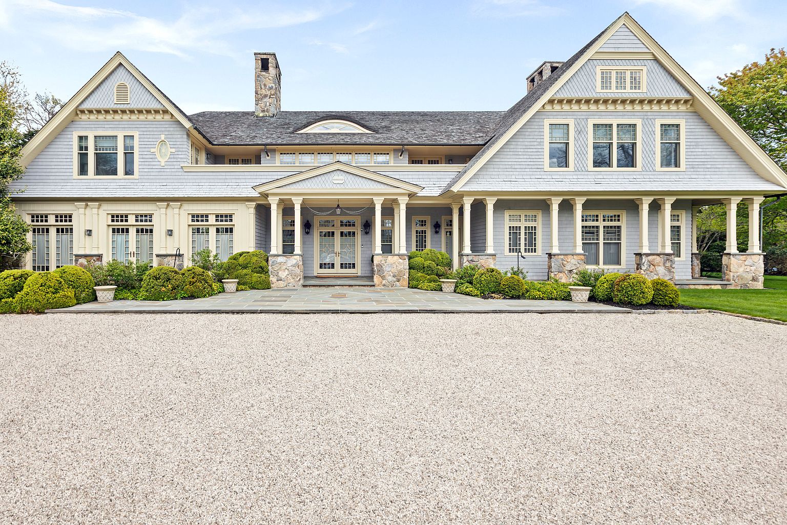 This grand, shingle-style residence features a symmetrical facade with light blue siding, cream-colored trim, and stone accents at the base of its columns. A wide, stone-paved walkway leads to a central covered entryway, framed by a gravel driveway that spans the foreground. The architecture is characterized by multiple gables, dormer windows, and a classic, elegant aesthetic that conveys a sense of luxury and curb appeal.