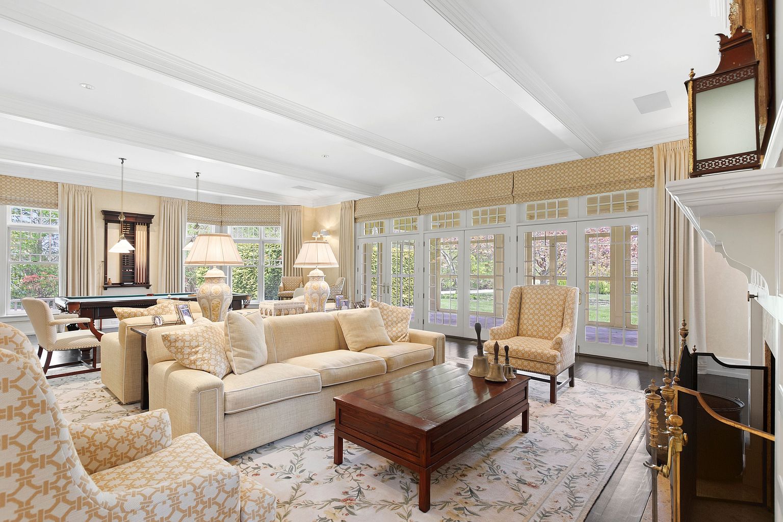 This spacious, light-filled living room features a neutral color palette with elegant patterned upholstery and a large, central coffee table. The room is anchored by a fireplace with a decorative mantel and is illuminated by soft, natural light streaming through a series of tall glass doors that open to the outdoors. A pool table is visible in the background, suggesting a versatile space designed for both relaxation and entertainment.