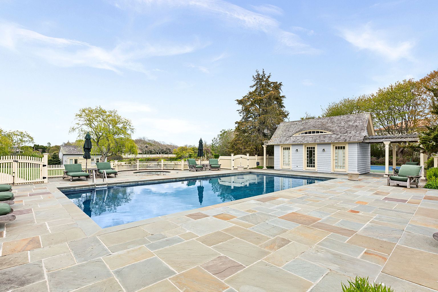 This expansive outdoor living area features a large, rectangular swimming pool surrounded by a spacious stone-paved patio. A charming, light-blue pool house with a shingled roof and pergola sits to the right, while multiple lounge chairs with green cushions are arranged around the pool deck. The scene is set against a backdrop of mature trees and a white picket fence, creating a serene and private atmosphere.
