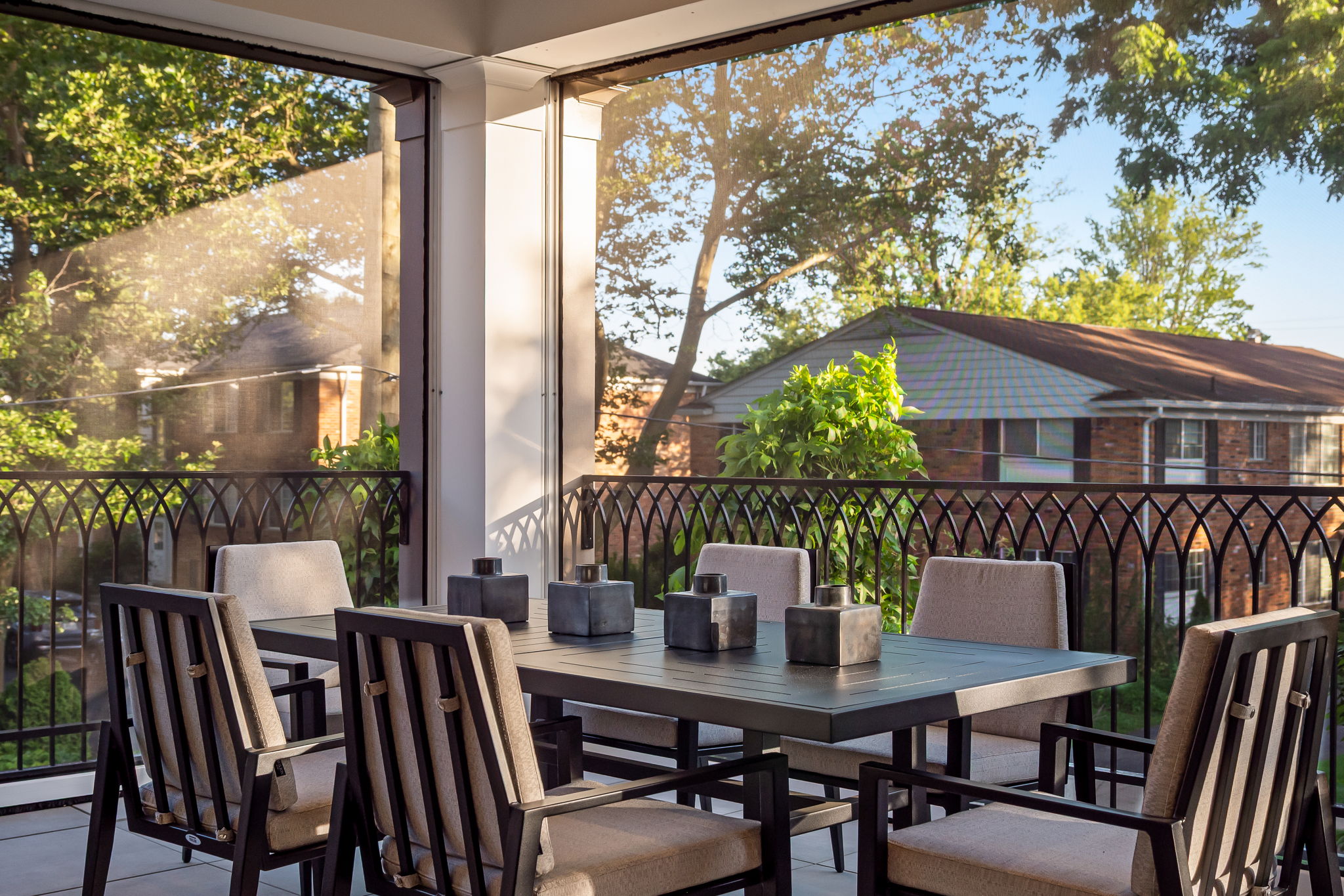 This image showcases a beautifully arranged outdoor dining area on a patio or balcony. The space features a dark-toned dining table surrounded by matching chairs with light-colored cushions, creating a sophisticated and inviting atmosphere. Decorative square vases adorn the table, and the wrought iron railing offers a glimpse of the surrounding greenery and neighborhood, enhancing the outdoor living experience.