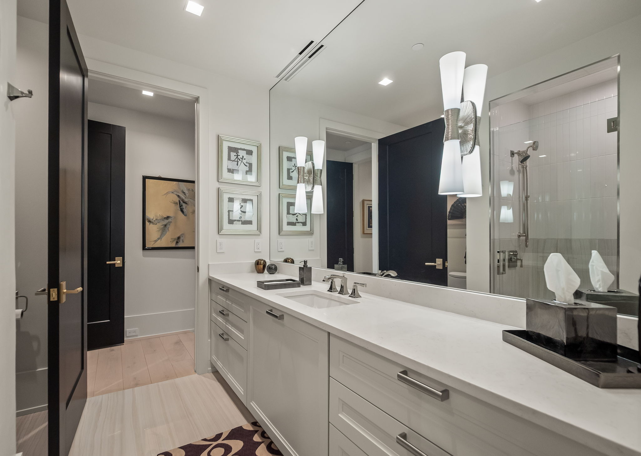 This is a primary bathroom featuring a long white vanity with ample storage, a large mirror, and modern lighting fixtures. The bathroom has a clean and contemporary design with a neutral color palette, complemented by dark-framed doors and artwork. A glass-enclosed shower is visible, adding to the luxurious feel of the space.