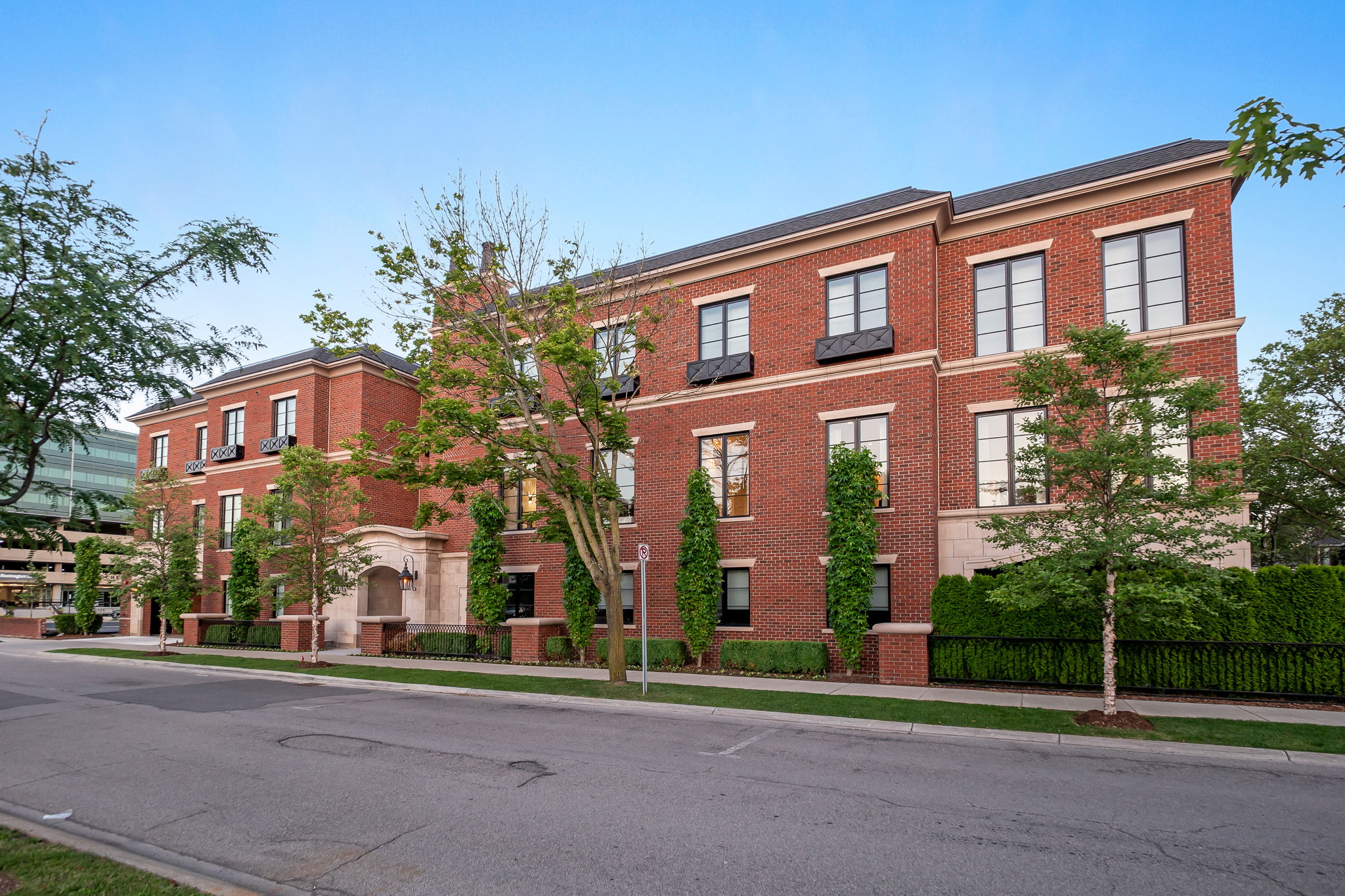 This is a front view of a stately brick building, likely a condominium or townhome complex, featuring symmetrical design elements and well-maintained landscaping. The building has multiple stories with evenly spaced windows, decorative window boxes, and a dark roof. A manicured lawn, trimmed hedges, and young trees line the front of the property, enhancing its curb appeal.