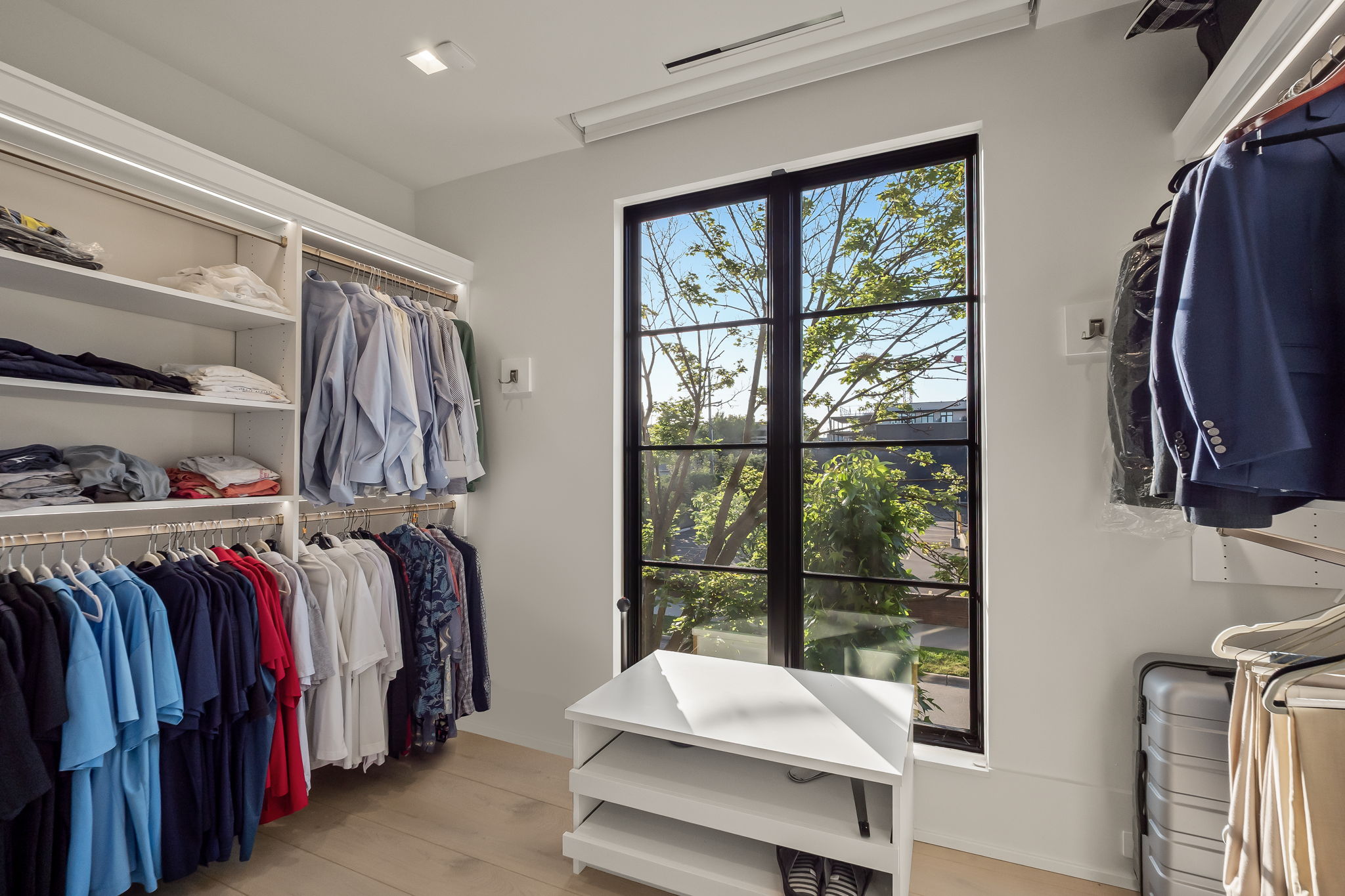 This is a well-organized walk-in closet featuring custom shelving and hanging rods. The closet is brightly lit, with natural light streaming in through a large window. A white storage bench sits beneath the window, and the overall impression is one of luxury and functionality.