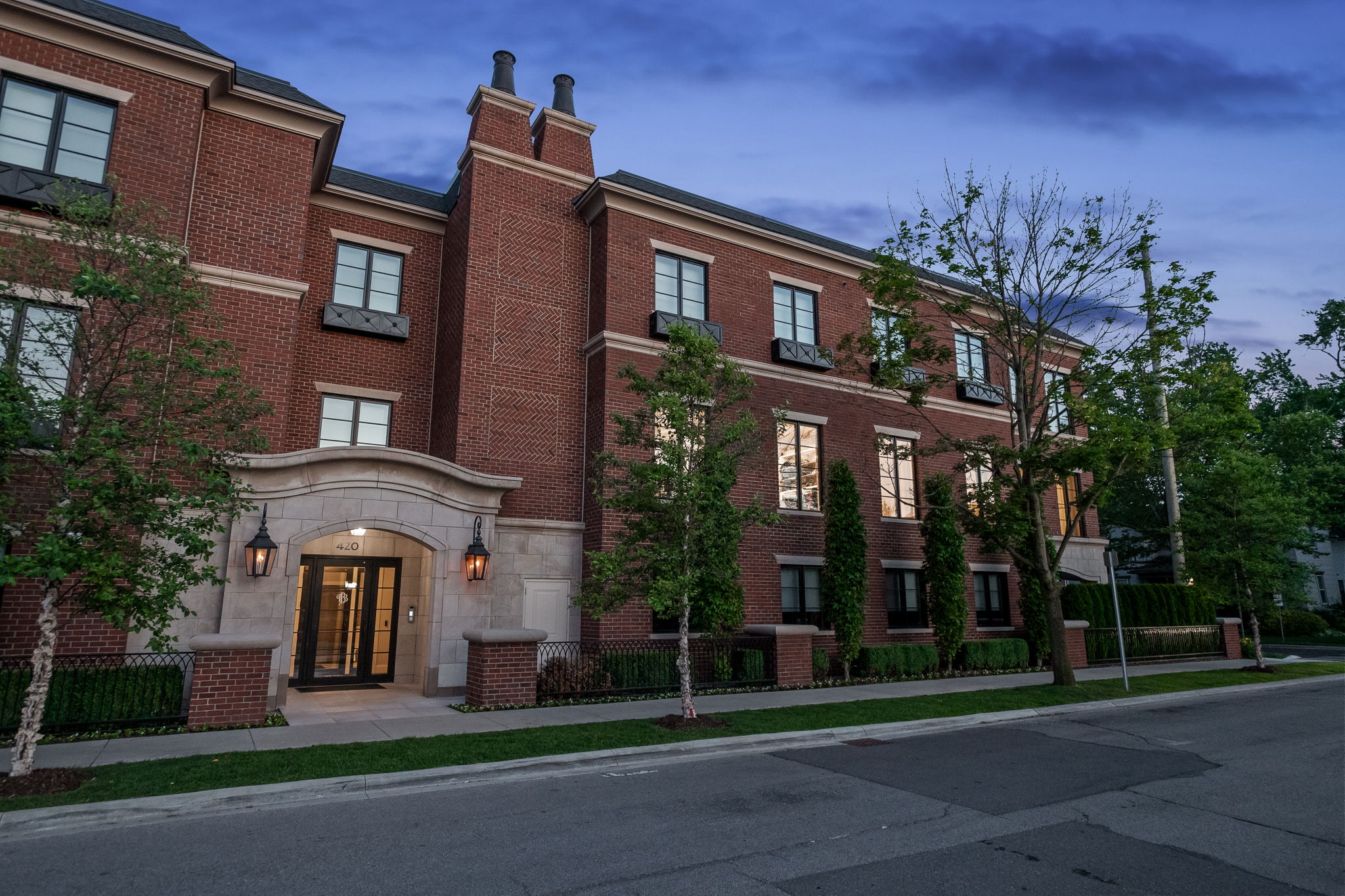 This is a front exterior view of a multi-story brick building with a prominent arched entryway marked with the number 420. The building features symmetrically placed windows with dark frames and decorative window boxes. Mature trees and manicured landscaping add to the property's curb appeal, while a paved street runs in front of the building, suggesting a well-maintained and accessible location.