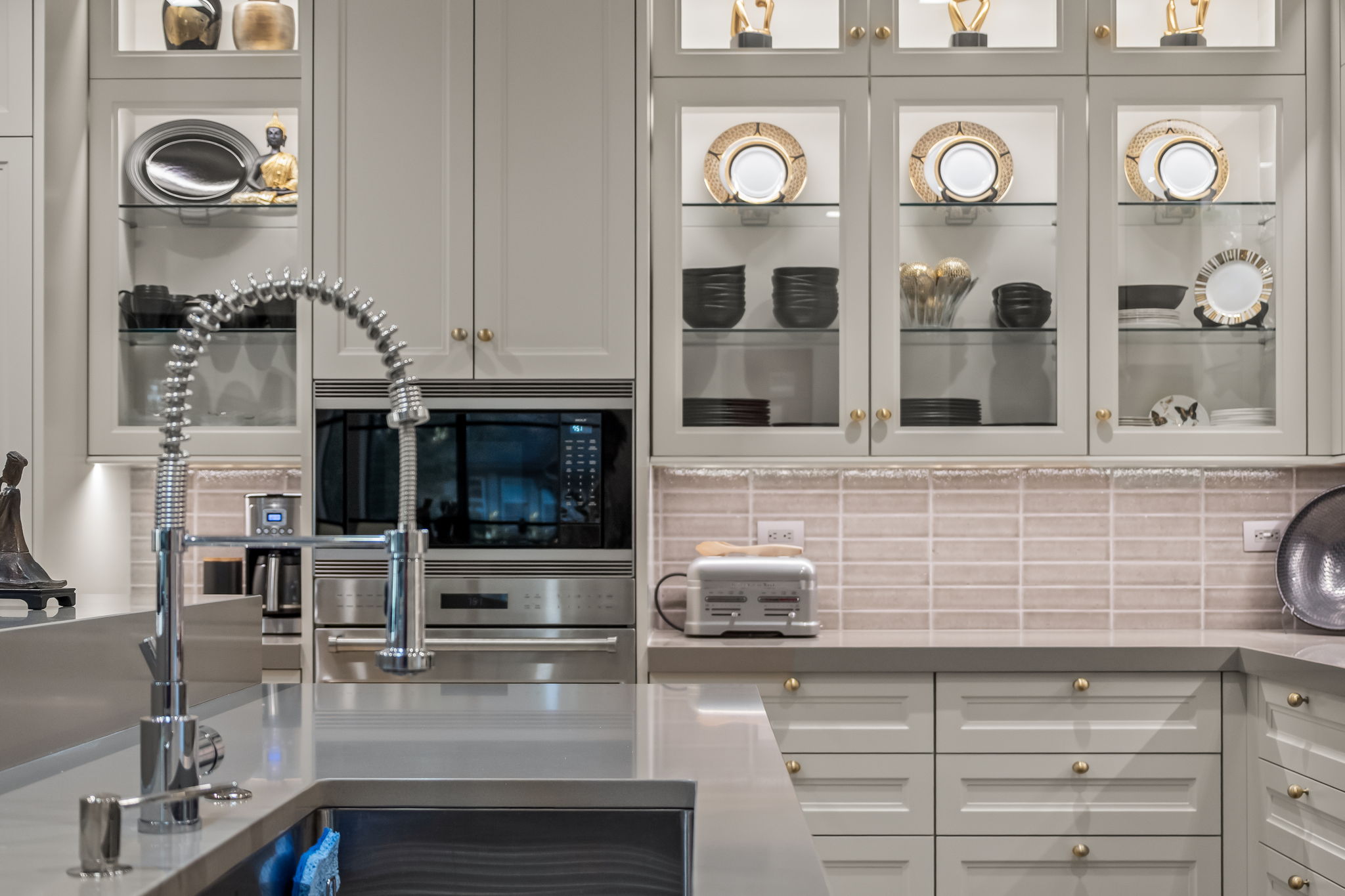 This is a detailed shot of a well-appointed kitchen featuring light gray cabinetry with gold hardware and glass-fronted display cabinets showcasing decorative dishware. Stainless steel appliances, including a built-in microwave and oven, are seamlessly integrated into the design. The kitchen also features a modern faucet and a light-colored backsplash, contributing to a clean and elegant aesthetic.