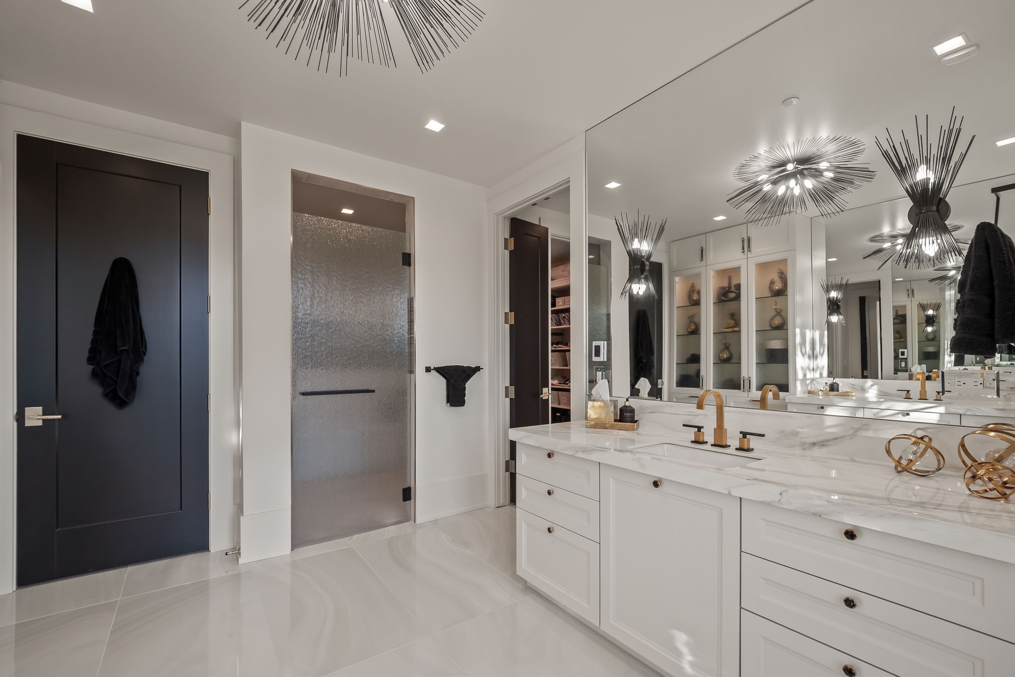 This is a luxurious primary bathroom featuring a double vanity with a white marble countertop and white cabinetry. A large mirror reflects the modern lighting fixtures and built-in storage cabinets. The bathroom also includes a glass-enclosed shower and a dark-colored door, contributing to a sophisticated and clean aesthetic.