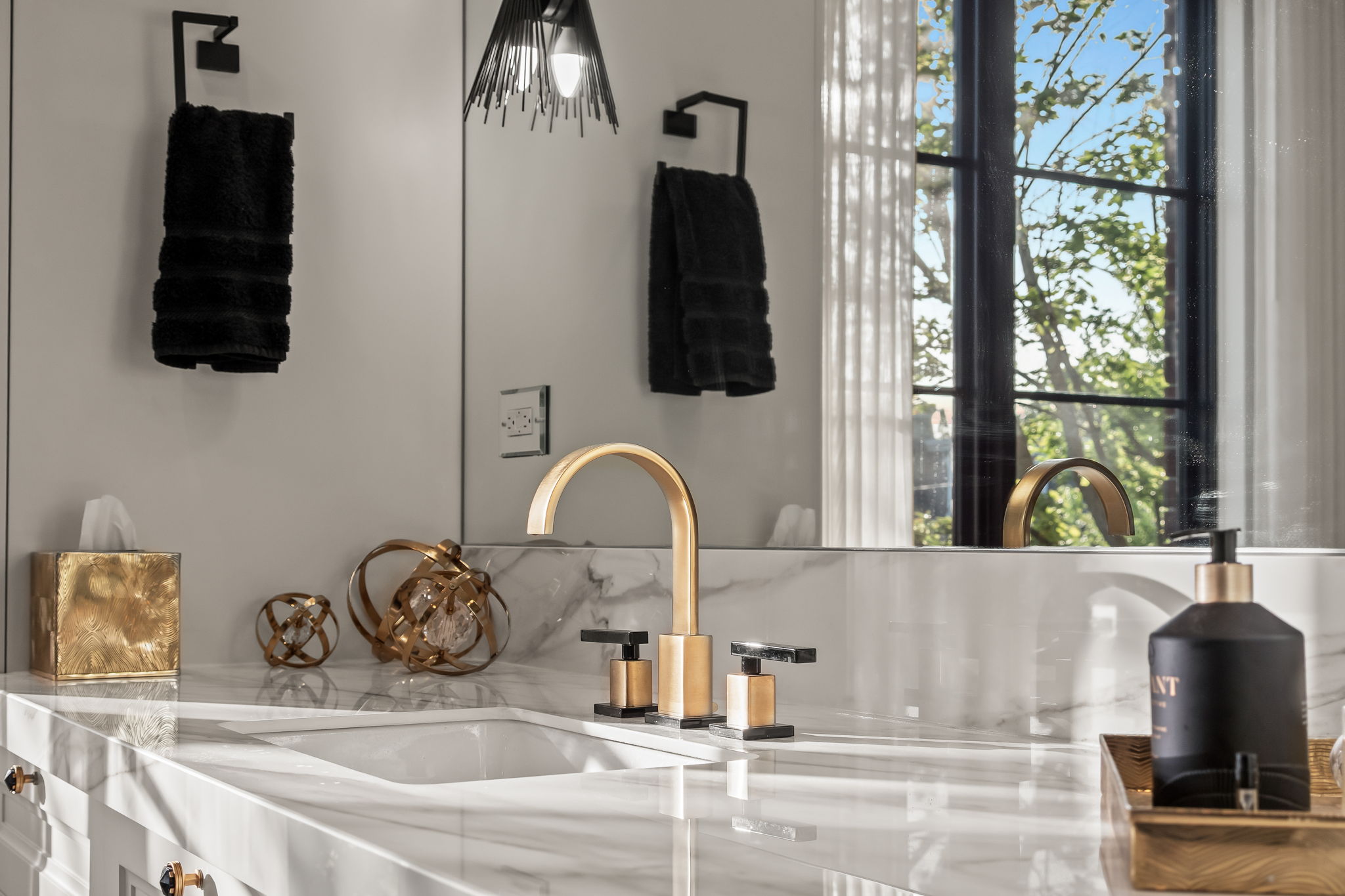 This is a detailed shot of a primary bathroom vanity area, showcasing a modern gold faucet with black handles set against a white marble countertop and backsplash. The vanity features white cabinetry with elegant hardware, and the mirror reflects black towel racks and a window view, creating a bright and luxurious atmosphere. The perspective is a medium shot, focusing on the vanity details.