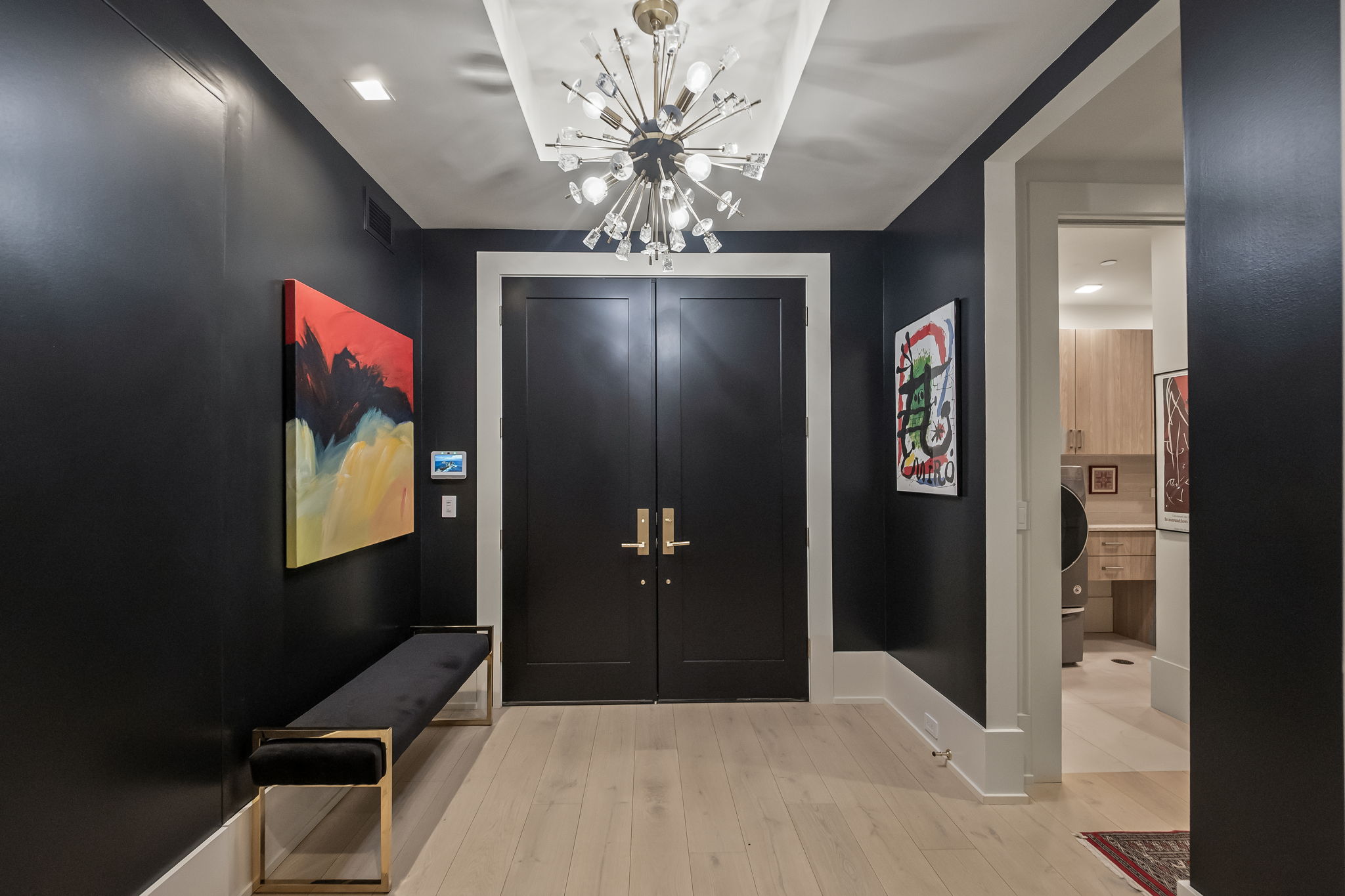 This is an interior shot of a modern hallway featuring dark walls, light wood floors, and a striking chandelier. A black bench with gold accents sits against the wall beneath an abstract painting, adding a touch of elegance. The hallway leads to a set of double doors and an open doorway, creating a sense of depth and flow.