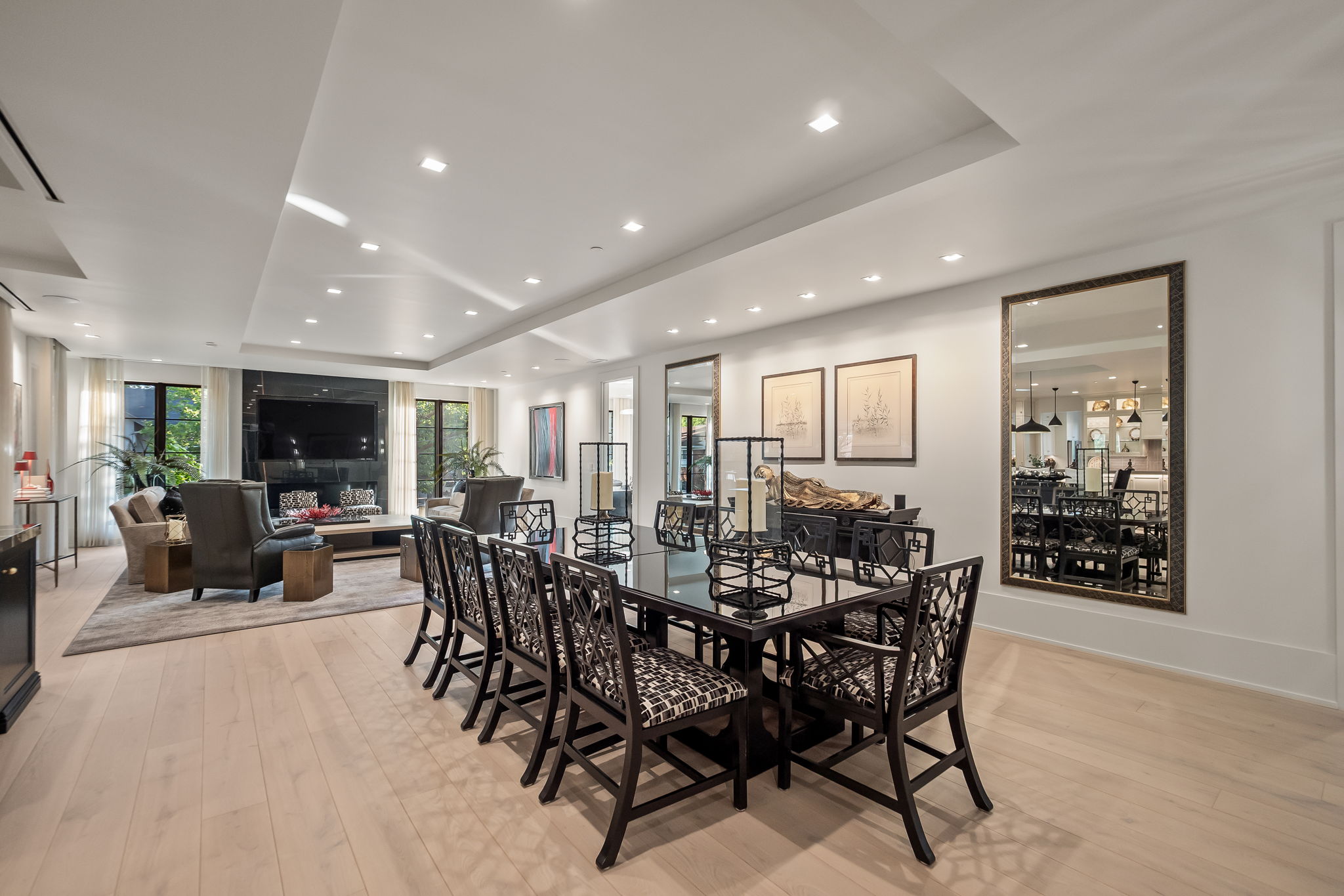 This interior shot showcases a spacious dining room that seamlessly blends into a living area. The dining area features a large, black glass-topped table surrounded by elegant, dark wood chairs with patterned upholstery. The room is well-lit with recessed lighting, and the light wood flooring adds warmth to the space. A large mirror on the wall reflects the dining area, creating a sense of depth and spaciousness.