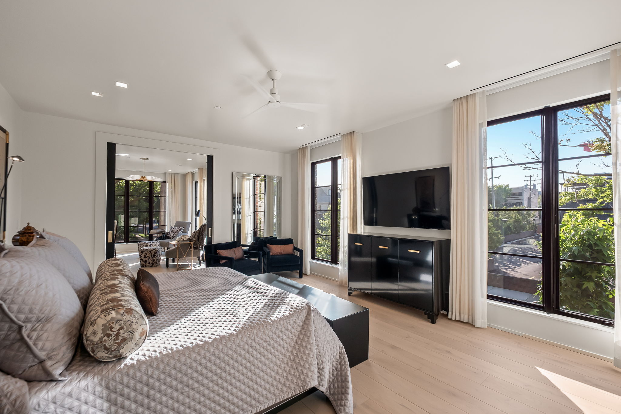 This primary bedroom features a large bed with a quilted coverlet and decorative pillows, a dark wood dresser with a large flat-screen TV above it, and two dark armchairs near a window. The room has light hardwood floors, white walls, and large windows that provide ample natural light. The overall impression is luxurious and serene.