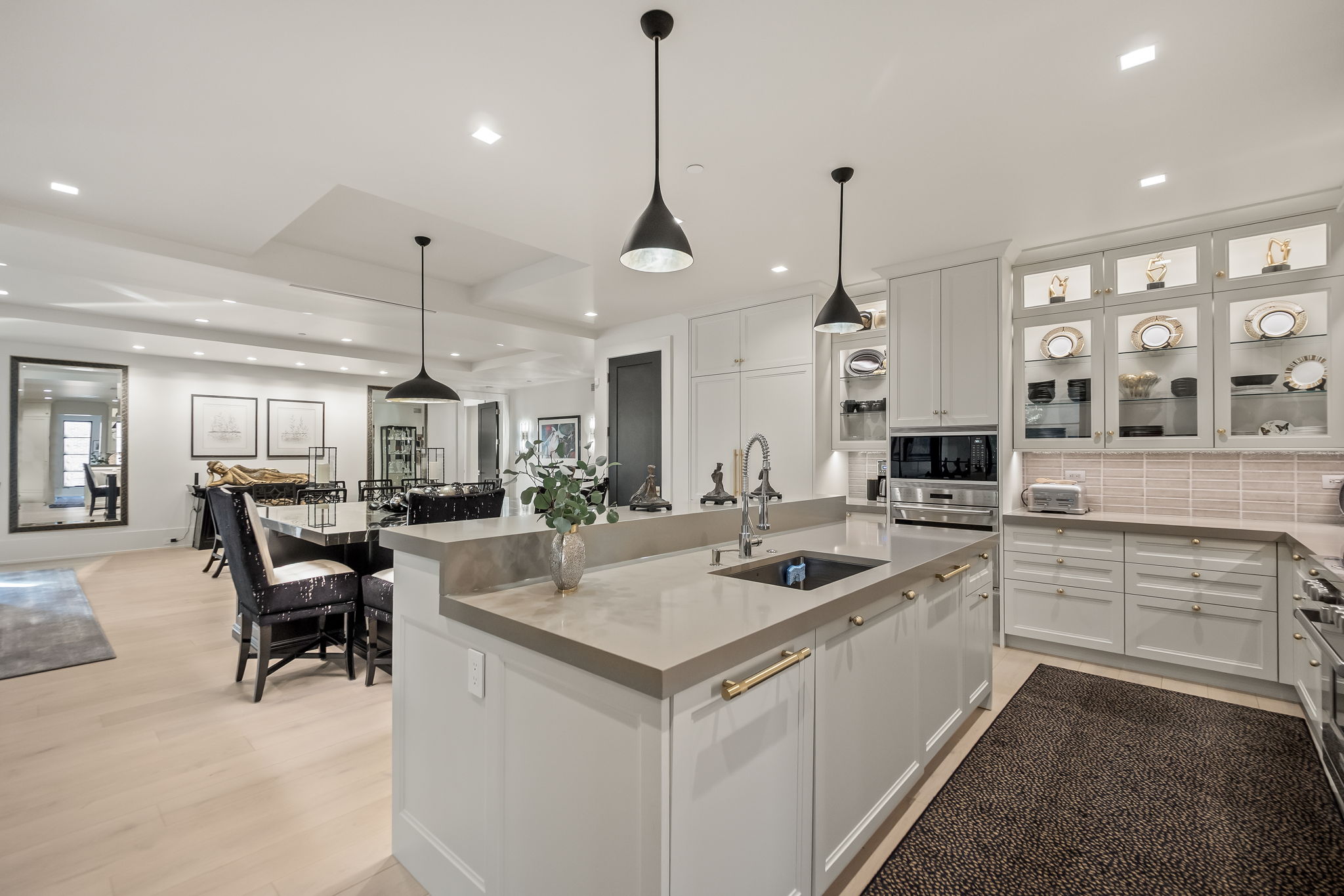 This is a bright and modern kitchen featuring a large island with a gray countertop and gold hardware. The kitchen is equipped with stainless steel appliances and custom cabinetry, including glass-front display cabinets. The open layout connects to a dining area, creating a seamless flow for entertaining.