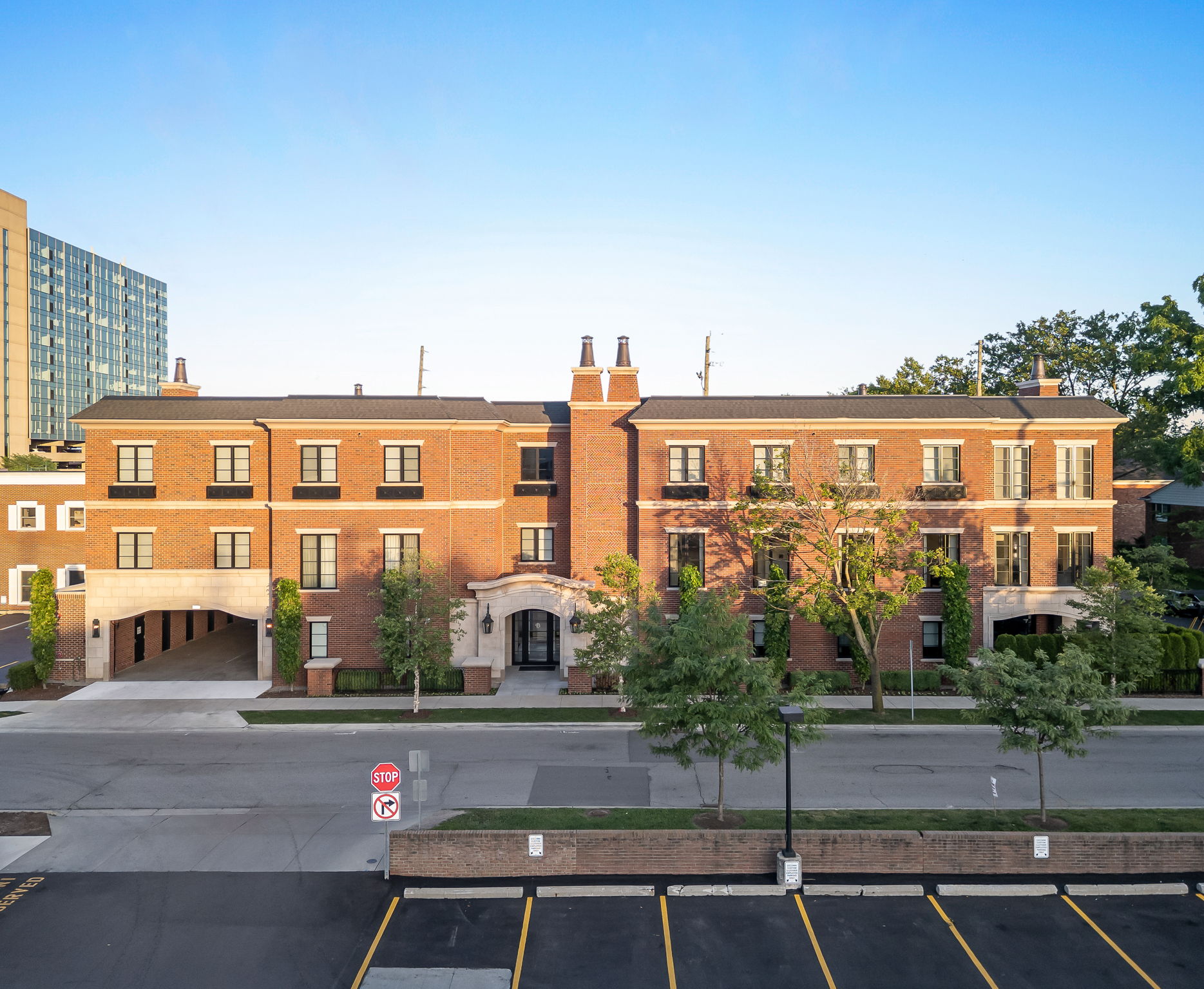 This aerial view showcases a well-maintained brick building with a symmetrical design and a central arched entryway. The building features multiple windows with dark frames, a dark roof, and decorative chimney elements. A parking lot is visible in the foreground, and mature trees and landscaping surround the property, adding to its curb appeal.