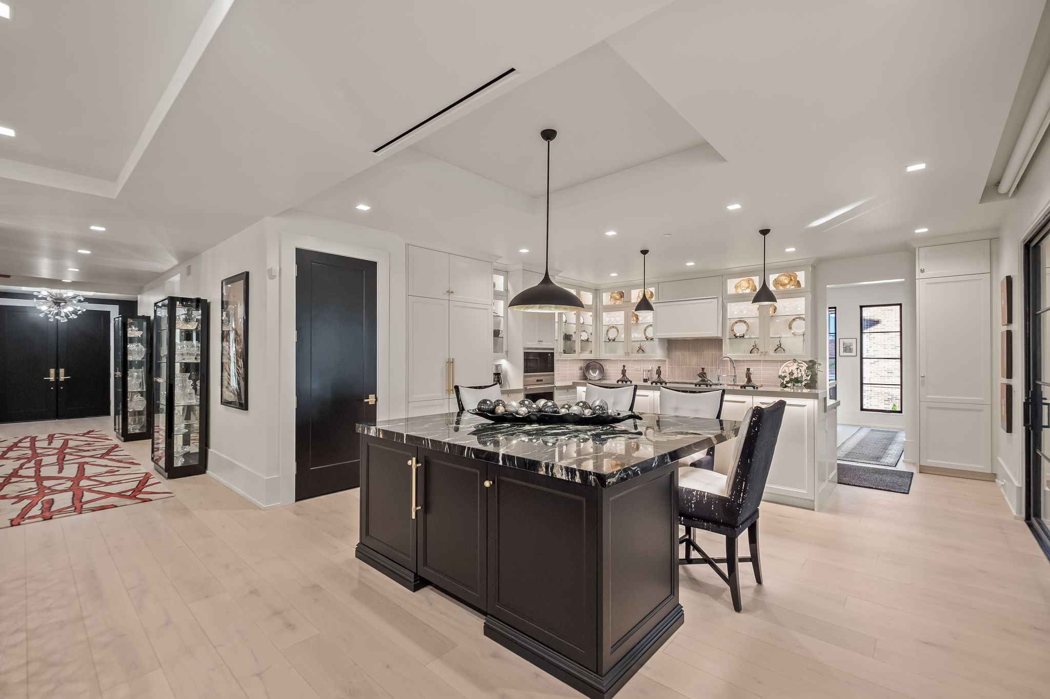 This is a bright and modern kitchen featuring a dark-colored island with a marble countertop as the focal point. The kitchen is equipped with white cabinetry, pendant lighting, and stainless steel appliances. The open layout connects to other living spaces, creating a seamless flow throughout the home.