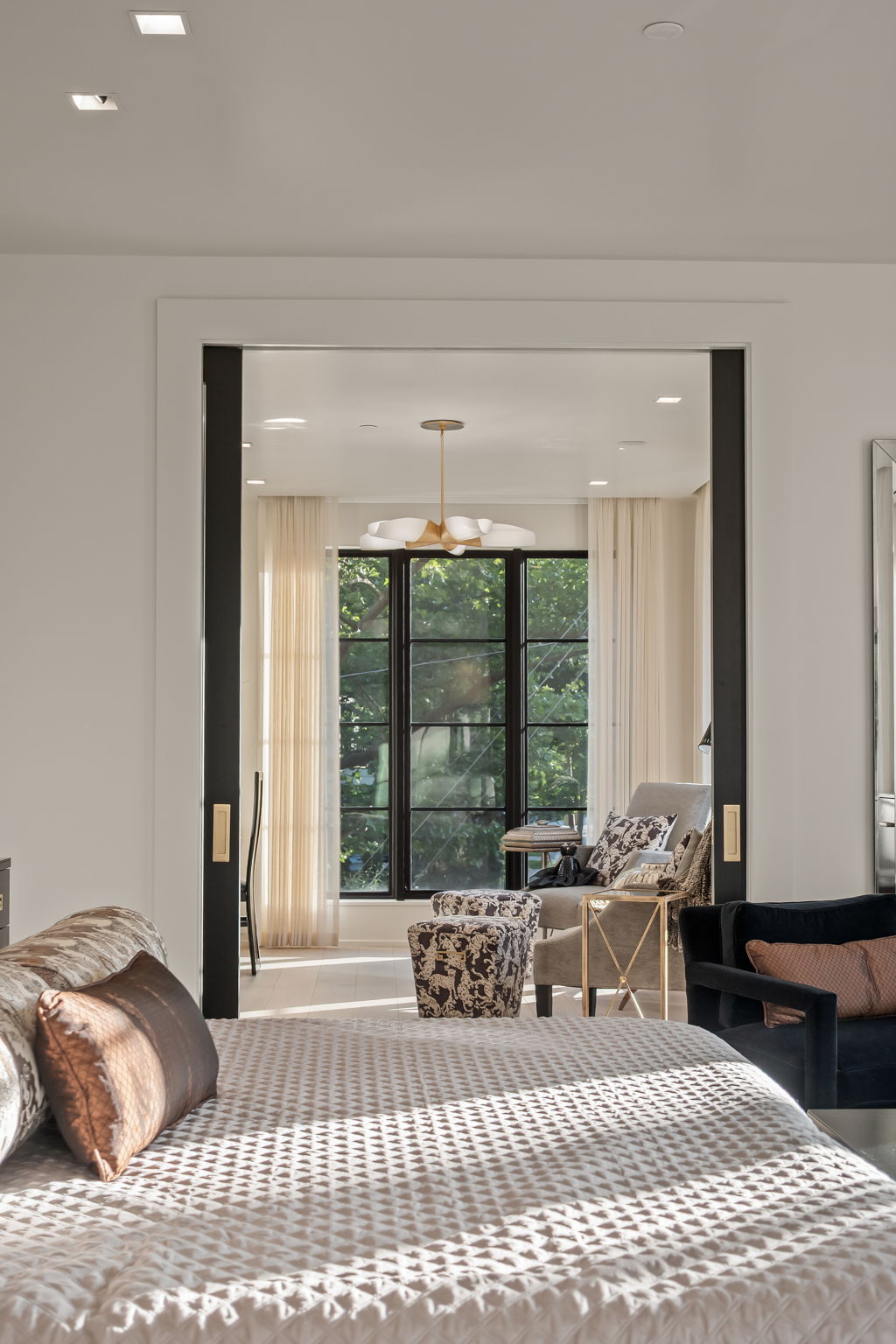 This image showcases a luxurious primary bedroom with a view into a sitting area. The bedroom features a textured bedspread and decorative pillows, while the sitting area beyond boasts a stylish chair, ottoman, and a modern chandelier. The black-framed doorway adds a touch of sophistication, creating an inviting and elegant space.