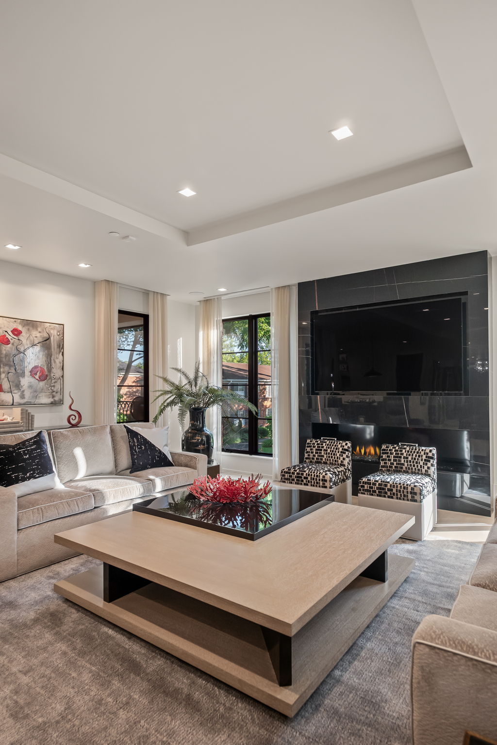 This is a well-lit living room featuring a large, modern coffee table as the focal point, surrounded by comfortable sofas and armchairs. A sleek, dark marble fireplace with a mounted television adds a touch of luxury, while large windows with sheer curtains provide natural light. The room exudes a sophisticated and inviting atmosphere.