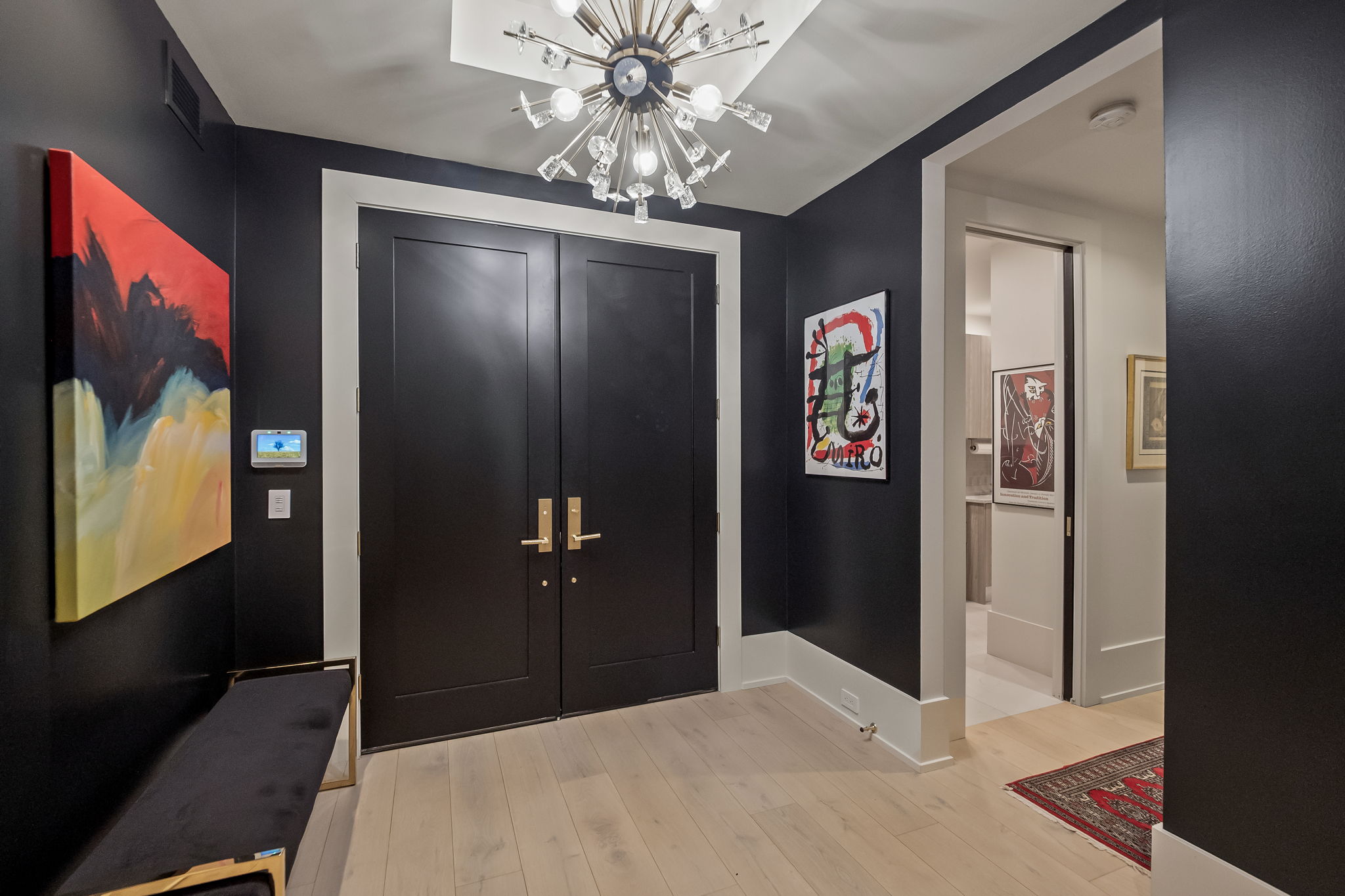 This is an interior shot of a modern hallway featuring dark walls contrasted by white trim and light wood flooring. A striking chandelier hangs from the ceiling, and artwork adorns the walls, adding a touch of sophistication. The hallway leads to another room, creating a sense of depth and flow within the home.