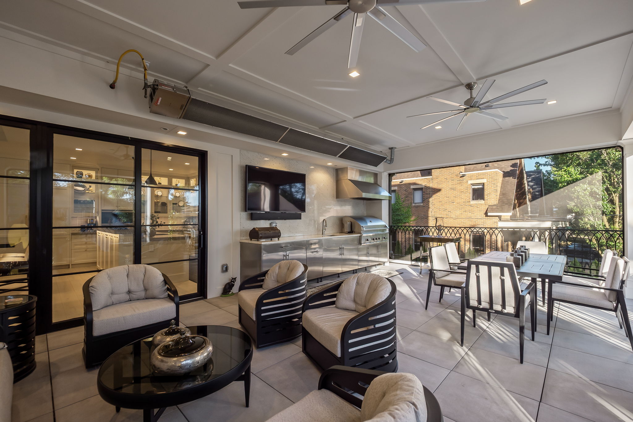 This image showcases a luxurious outdoor patio area, featuring comfortable lounge seating arranged around a modern coffee table. An outdoor kitchen with stainless steel appliances and a built-in grill adds functionality, while a dining table set provides space for outdoor meals. The space is well-lit and appears to be designed for relaxation and entertainment.