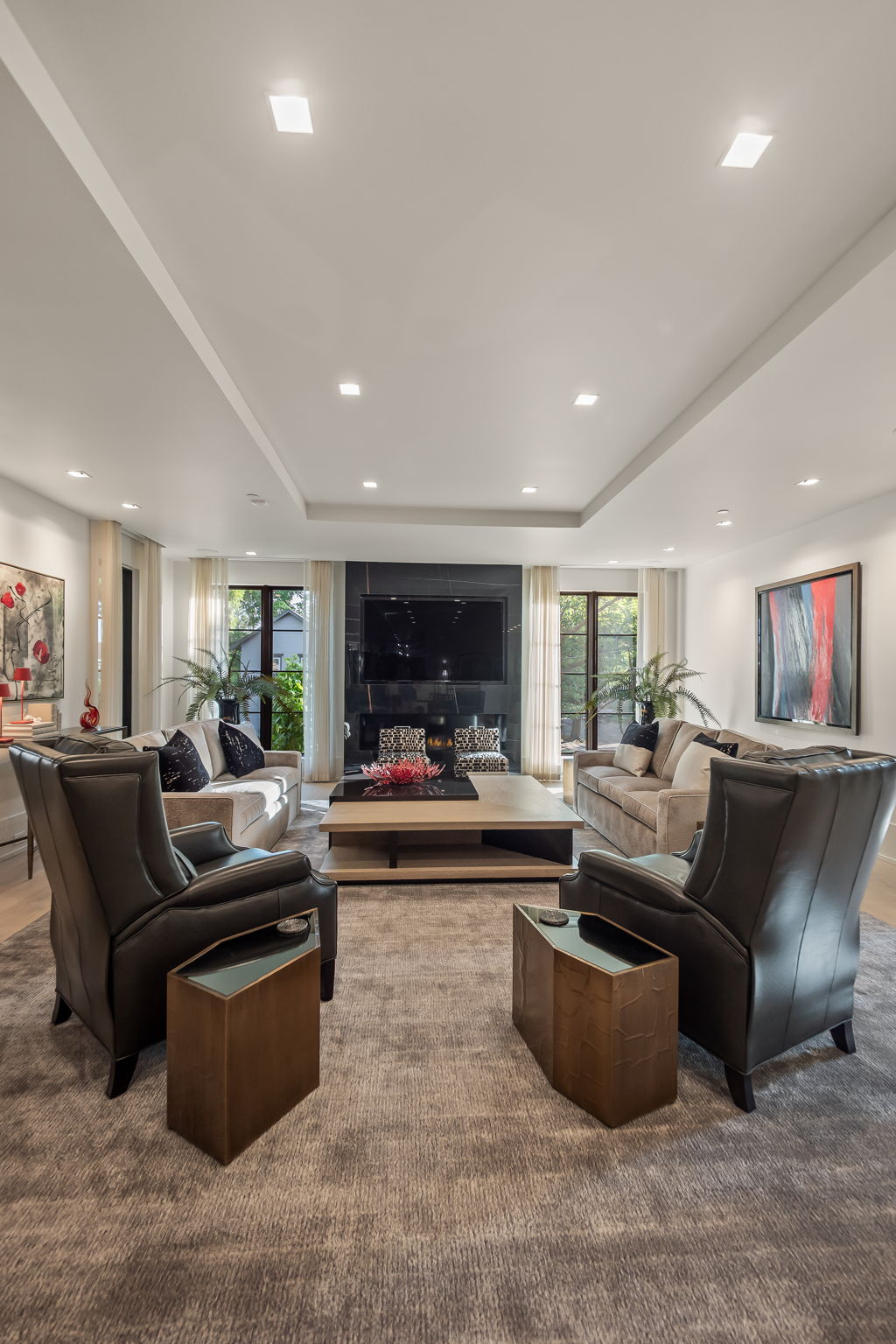This is a well-appointed living room featuring a neutral color palette with pops of black and red in the artwork. The room is anchored by a large gray rug and includes two sofas, two armchairs, and a substantial coffee table. A modern fireplace with a black marble surround and a large television above it serve as a focal point, flanked by windows with sheer curtains.