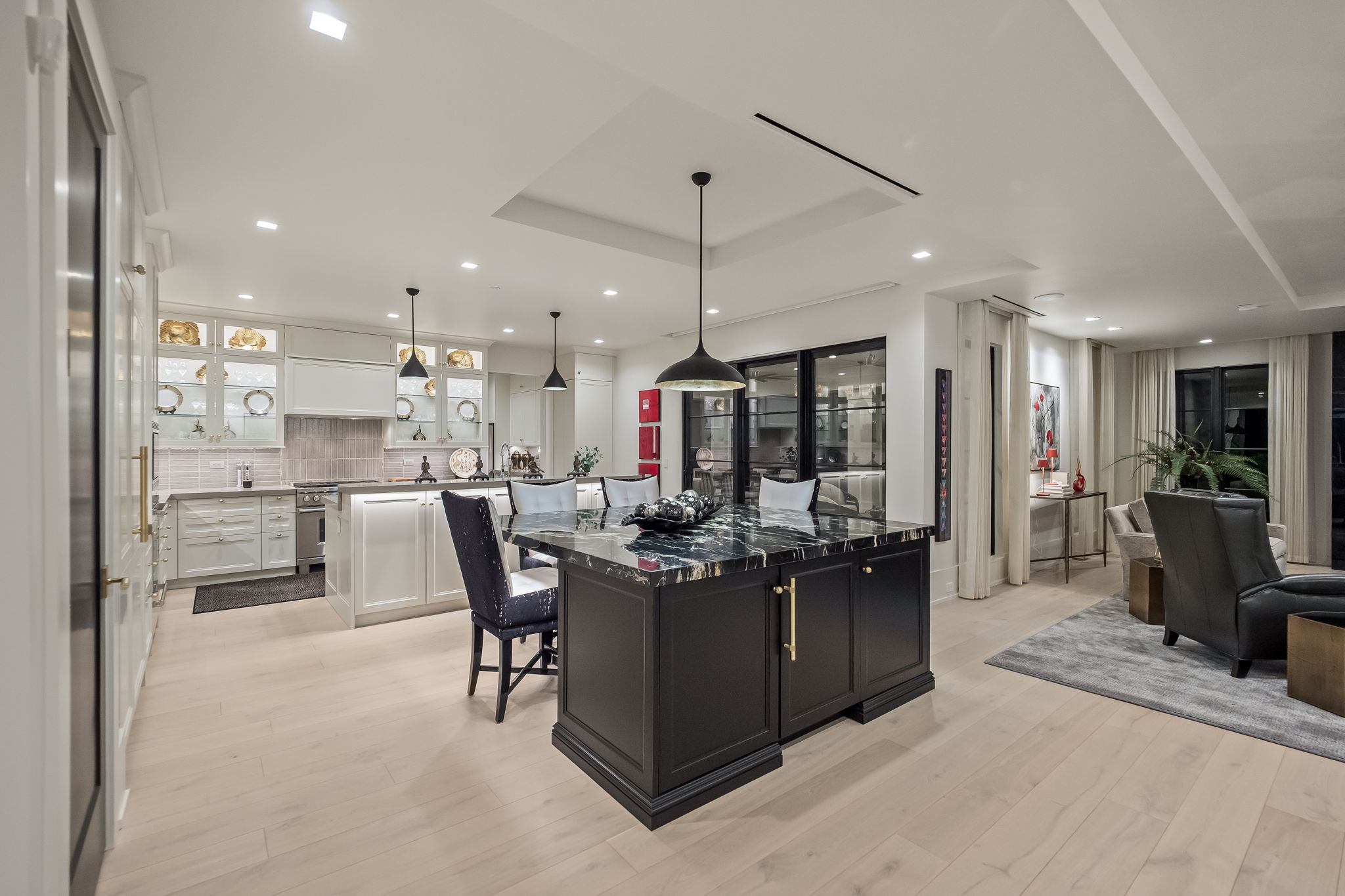 This is a bright and modern kitchen featuring a large dark island with a marble countertop and gold hardware, complemented by white cabinetry and stainless steel appliances. The open-concept design flows into a living area, creating a spacious and inviting atmosphere. Pendant lights hang above the island, adding a touch of elegance to the space.