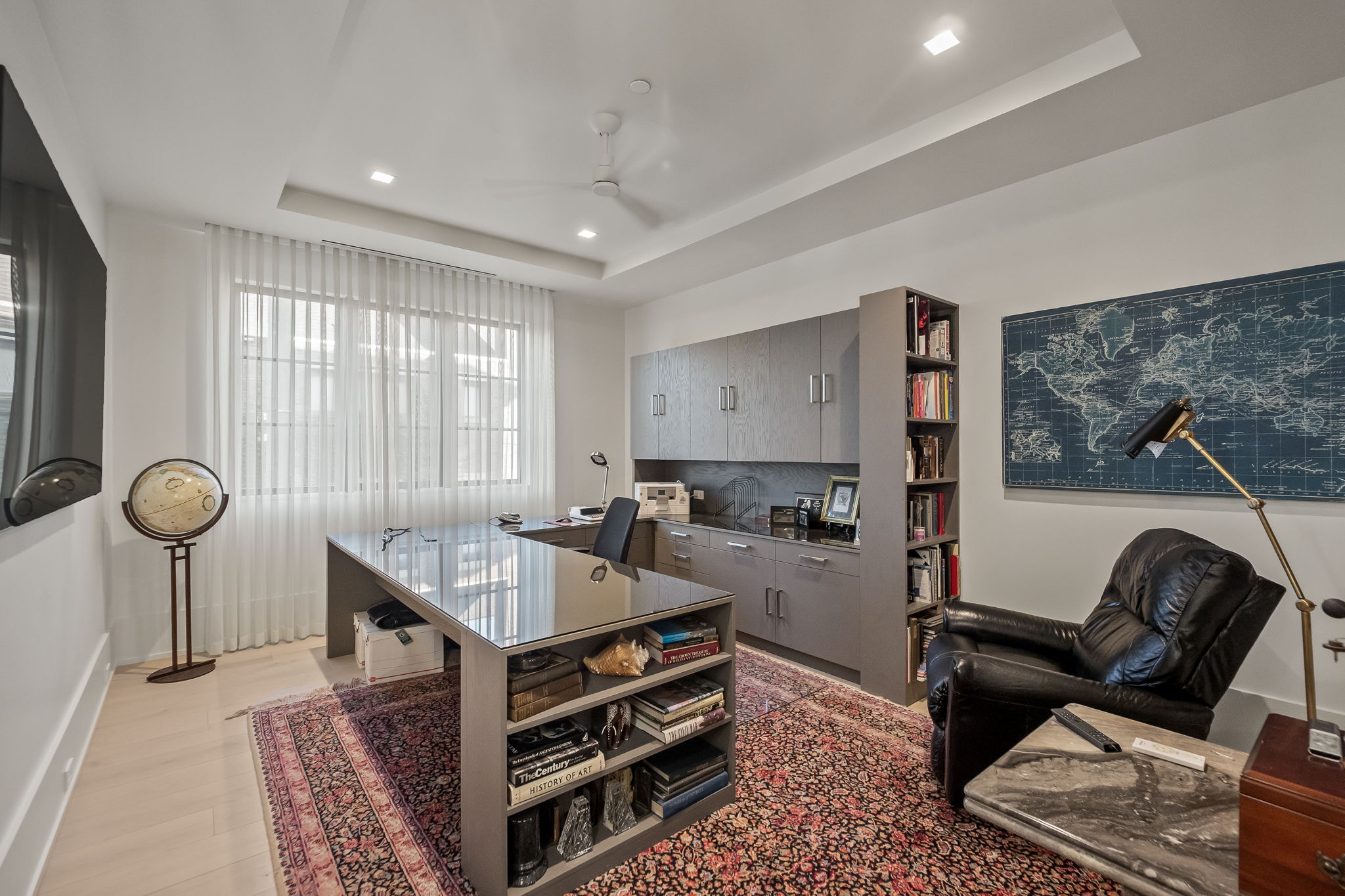 This is a well-appointed home office featuring a large glass-topped desk with built-in shelving, a comfortable leather armchair, and custom gray cabinetry with integrated shelving. A world map hangs on the wall, complemented by a stylish desk lamp, and a patterned rug adds warmth to the space. The room is bright and airy, with natural light filtering through sheer curtains.