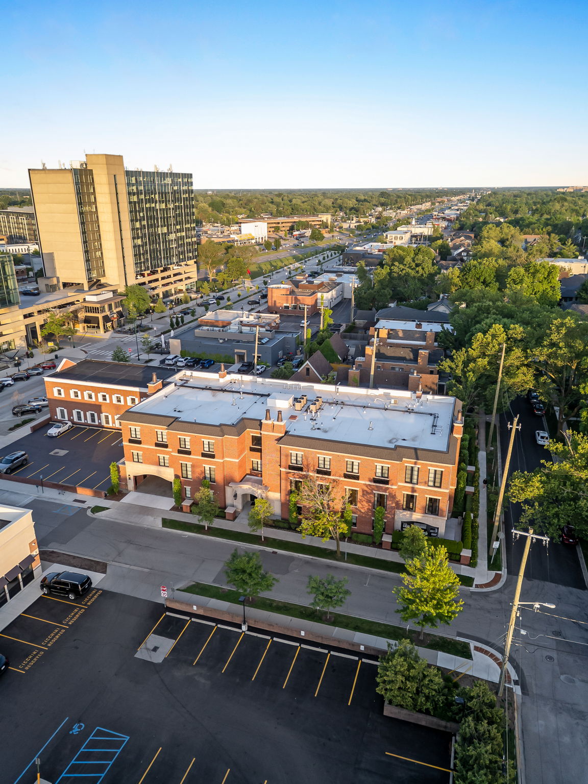 This aerial view showcases a well-maintained brick building with a light-colored roof, surrounded by lush greenery and a neatly organized parking lot. The building's architecture features symmetrical window placement and a central archway, suggesting a sophisticated and inviting atmosphere. The surrounding cityscape includes modern high-rise buildings and a tree-lined street, indicating a desirable urban location.