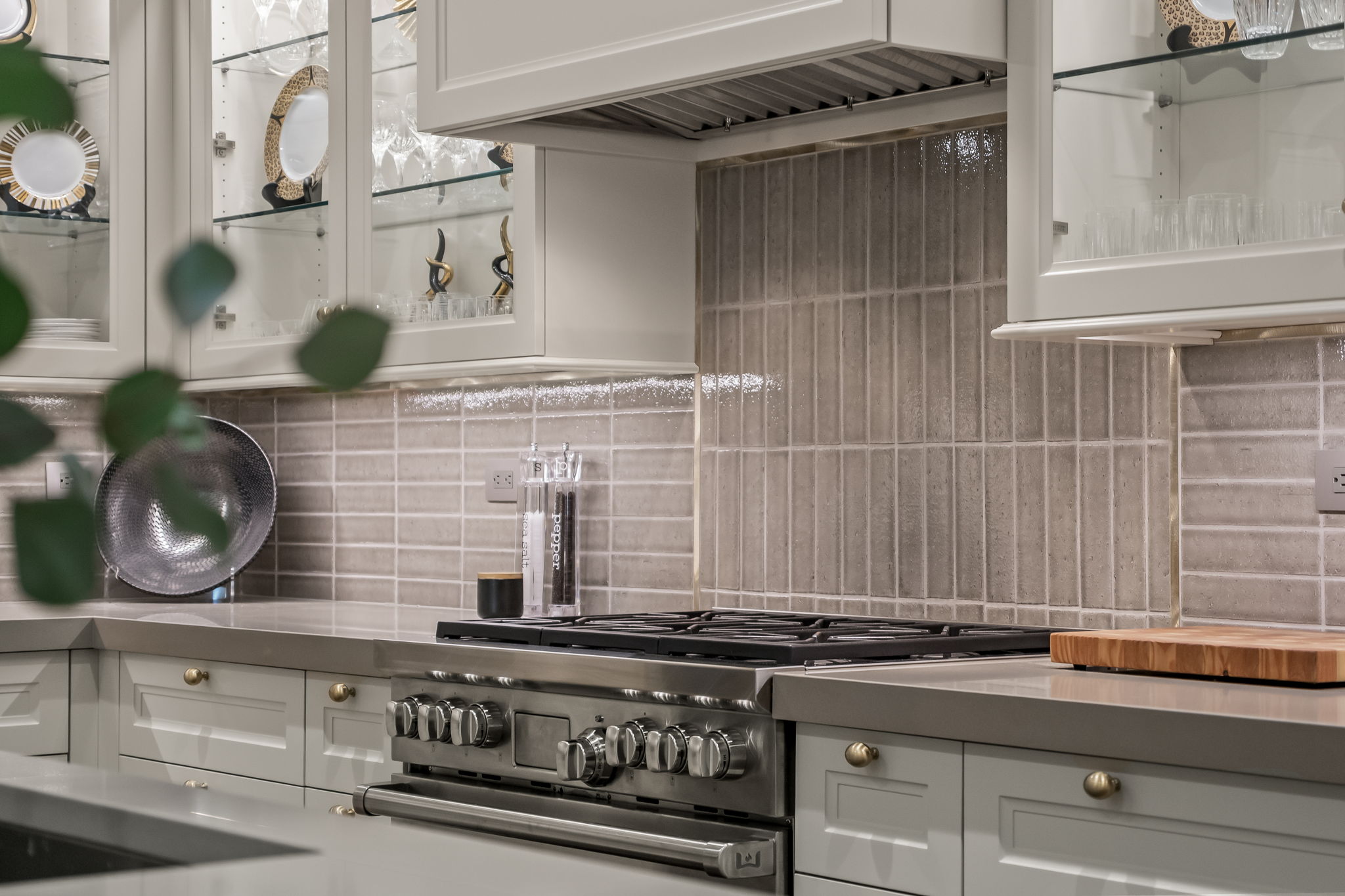This is a detailed shot of a kitchen featuring a stainless steel gas range with multiple knobs and burners, set against a backdrop of light gray, horizontally-laid tile backsplash. The countertops are a matching light gray, and the cabinets are a classic white with brass knobs. Glass-front cabinets display decorative plates and glassware, adding a touch of elegance.