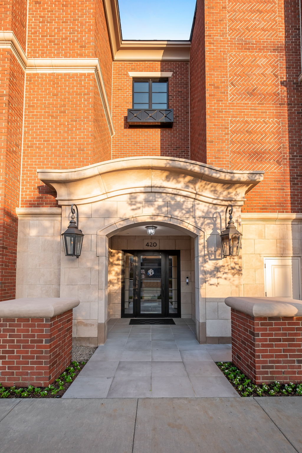 This is a well-lit, inviting entryway to a brick building. The entrance features a stone archway with elegant lighting fixtures on either side, leading to a glass door with a decorative emblem. The walkway is paved with large stone tiles, flanked by brick planters with greenery, creating a sophisticated and welcoming first impression.