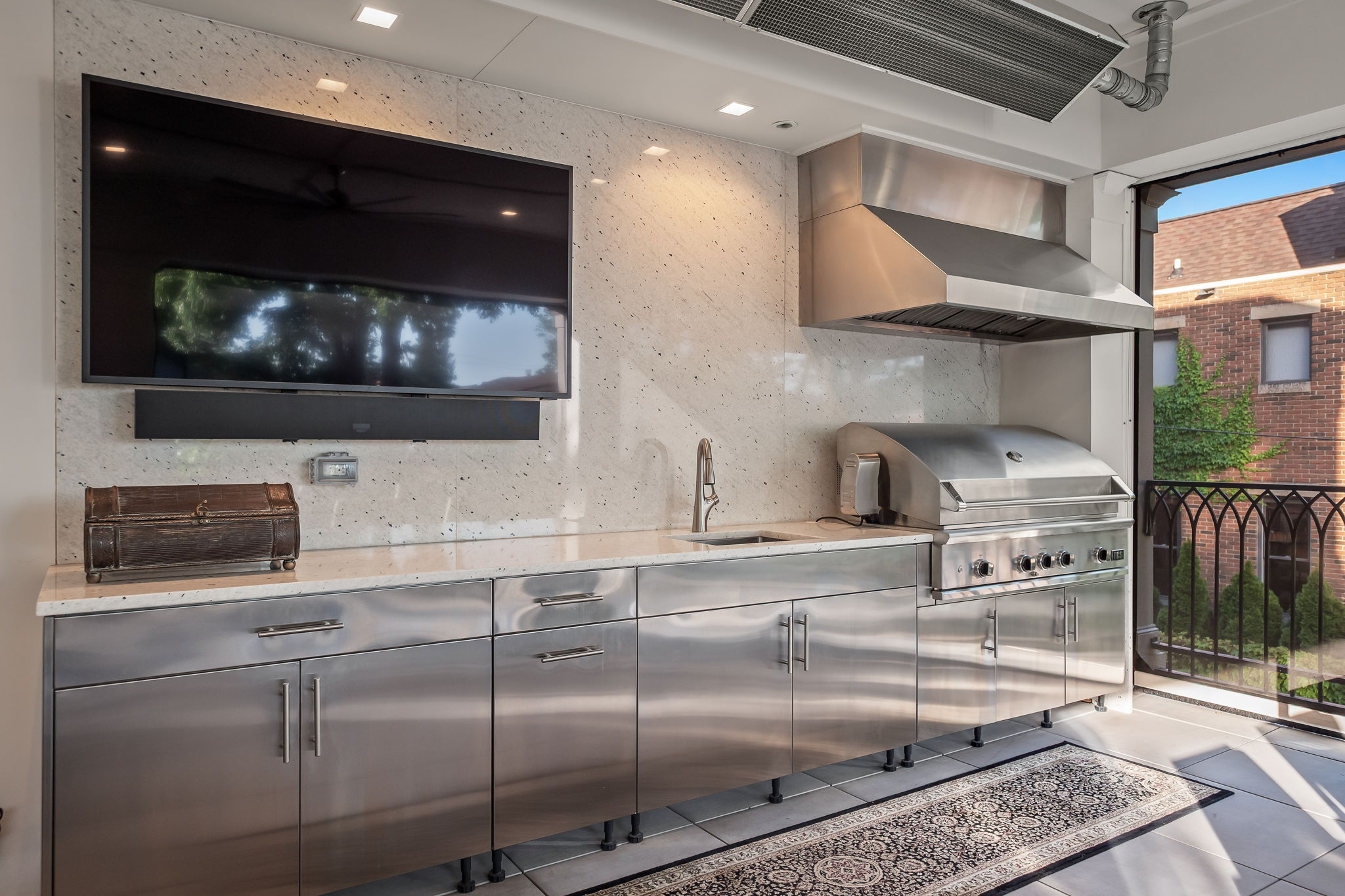This image showcases an outdoor kitchen area featuring stainless steel cabinetry, countertops, and a built-in grill with a hood. A large television is mounted on the wall, and a decorative chest sits on the counter. The space opens to an outdoor area with a view of a brick building and greenery, creating a seamless indoor-outdoor living experience.