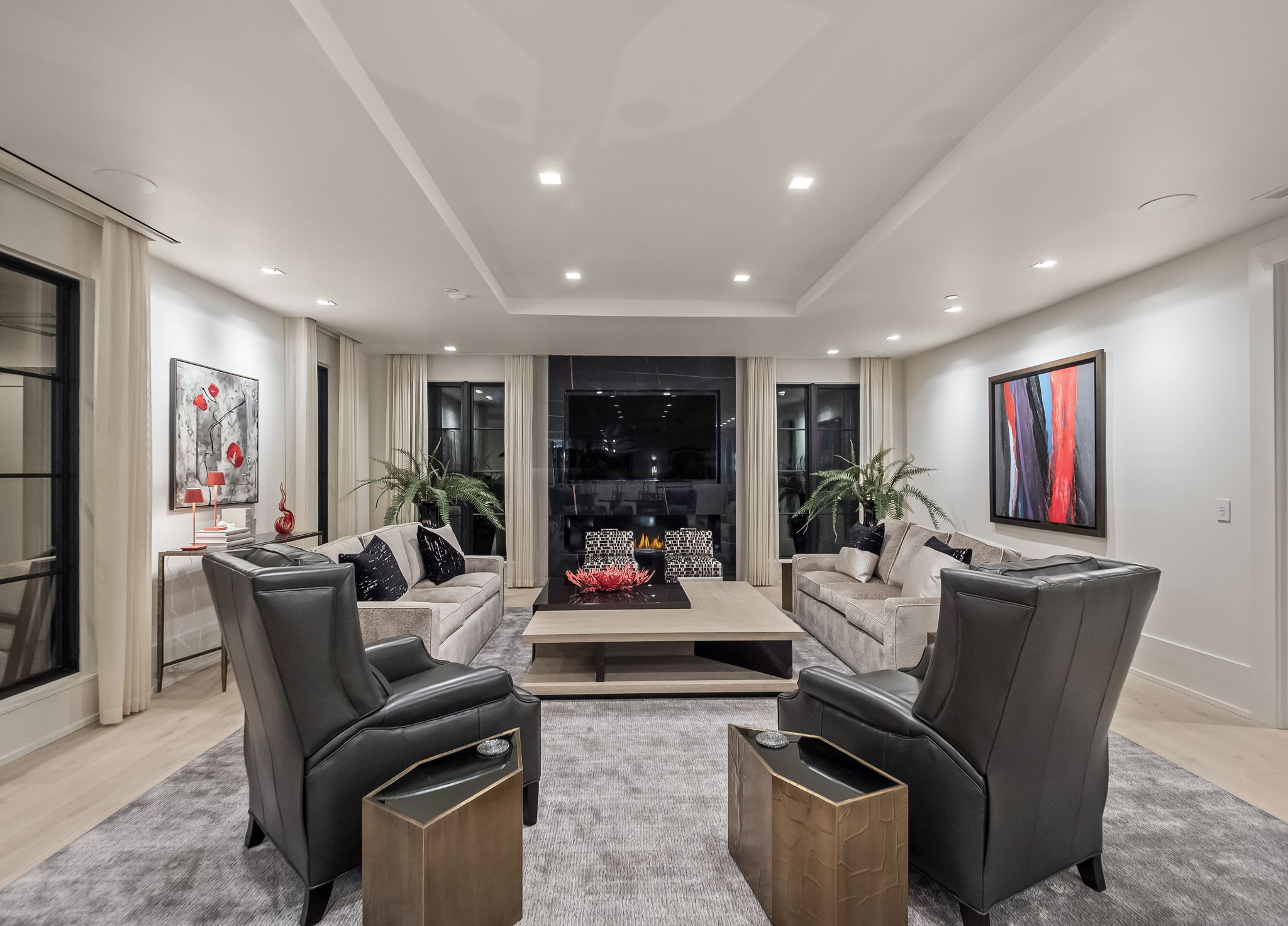 This is a luxurious living room featuring a symmetrical layout with two sofas facing each other, flanked by black leather armchairs. A large, modern coffee table sits in the center on a gray rug, and a fireplace with a television above it serves as a focal point. The room is well-lit with recessed lighting and natural light from the windows, creating a sophisticated and inviting atmosphere.