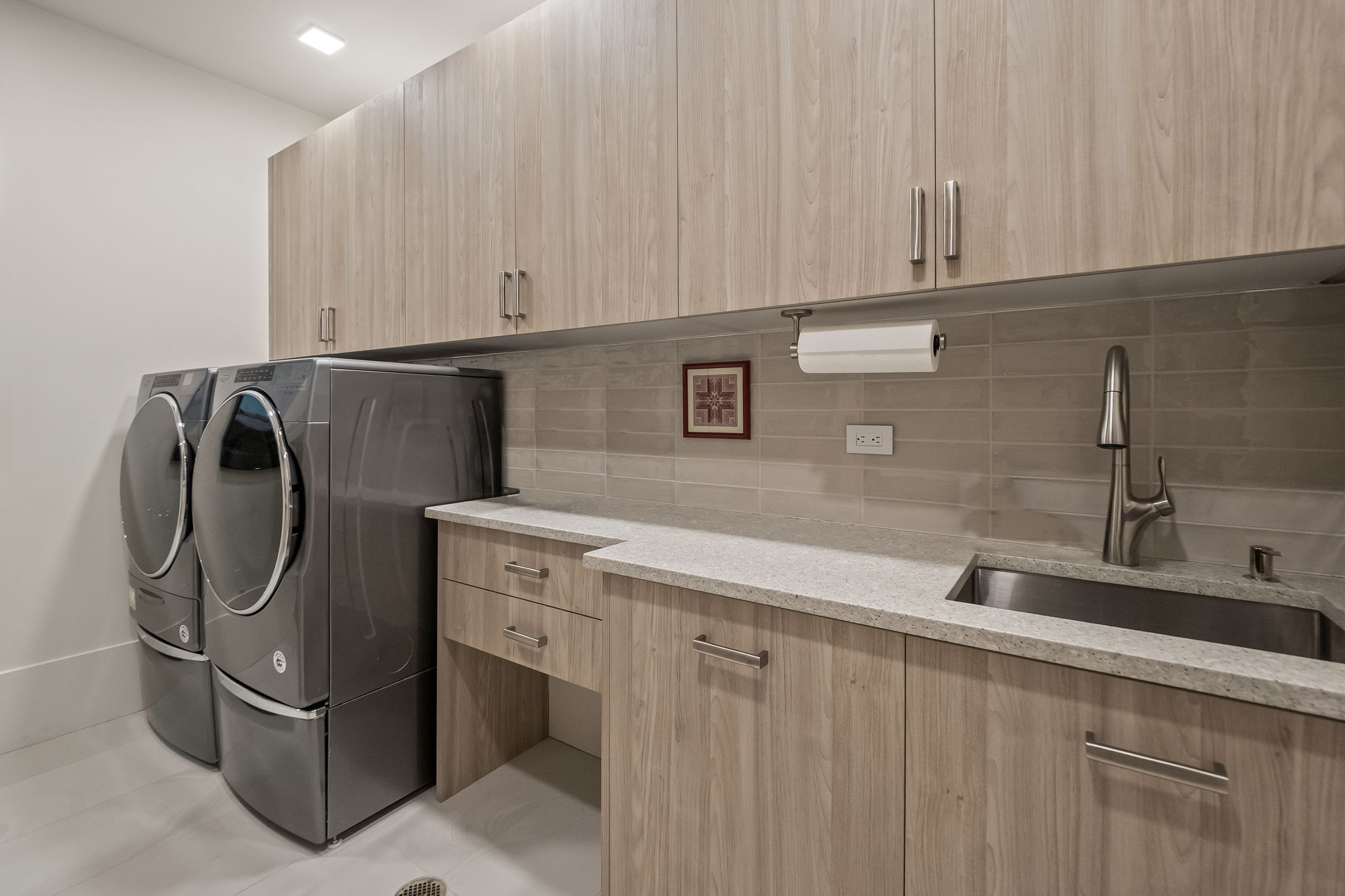 This is a well-organized laundry room featuring a front-loading washer and dryer set in a sleek gray finish. The room includes light wood-grain cabinets above a countertop with a stainless steel sink and modern faucet. The neutral color palette and clean lines create a functional and aesthetically pleasing space.