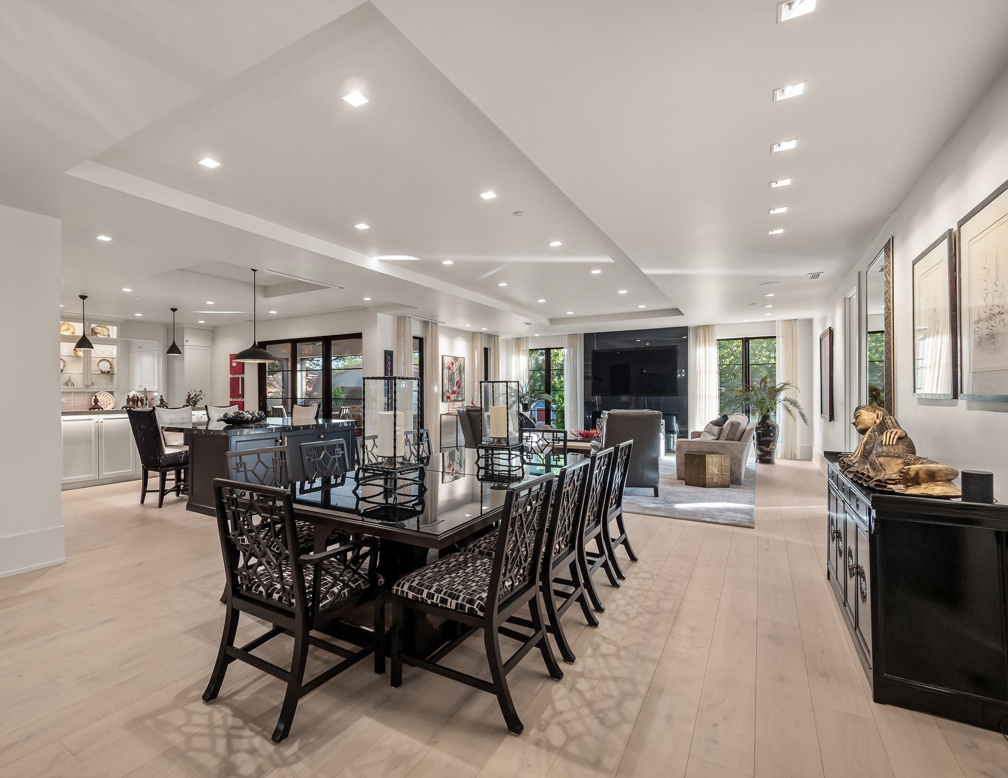 This is an interior shot of a dining room that seamlessly transitions into a living room and kitchen area. The dining room features a large, dark-colored dining table with patterned chairs, while the living room offers comfortable seating and natural light through large windows. The overall impression is one of spaciousness and modern elegance, perfect for entertaining and comfortable living.