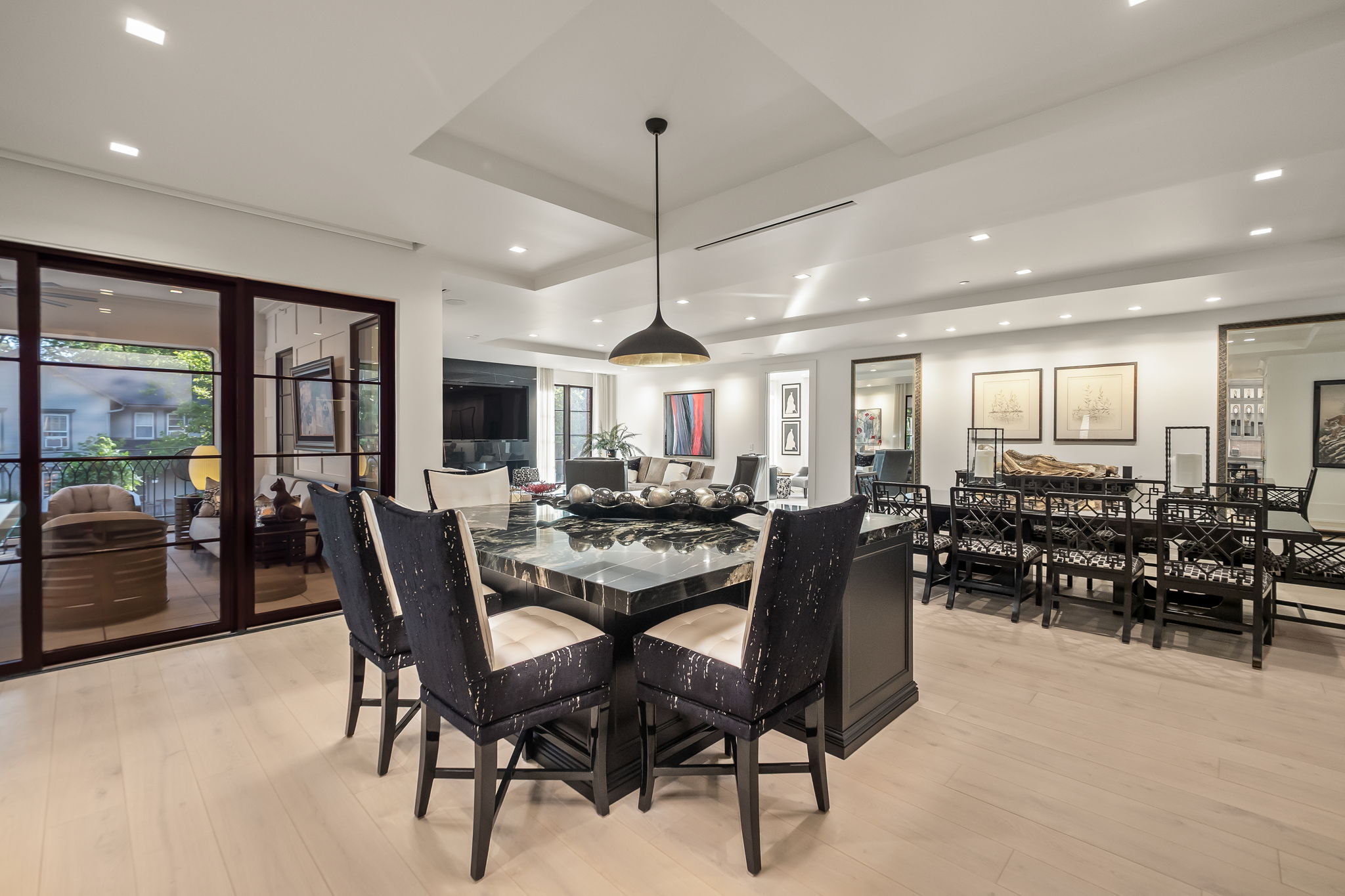 This is an interior shot of a dining room featuring a large, square, black marble dining table with seating for six. The room is well-lit with recessed lighting and a modern pendant light fixture hanging above the table. The space opens to a living area and has large sliding glass doors leading to an outdoor space, creating an open and airy feel.