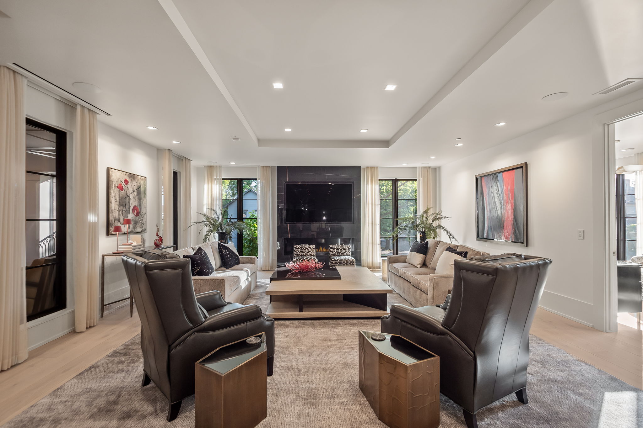 This is a luxurious living room featuring two sofas facing each other with a modern coffee table in between. Two leather armchairs are in the foreground, flanking the sofas. The room has a neutral color palette with black accents, including a fireplace and window frames, and the space is well-lit with recessed lighting and natural light from the windows.