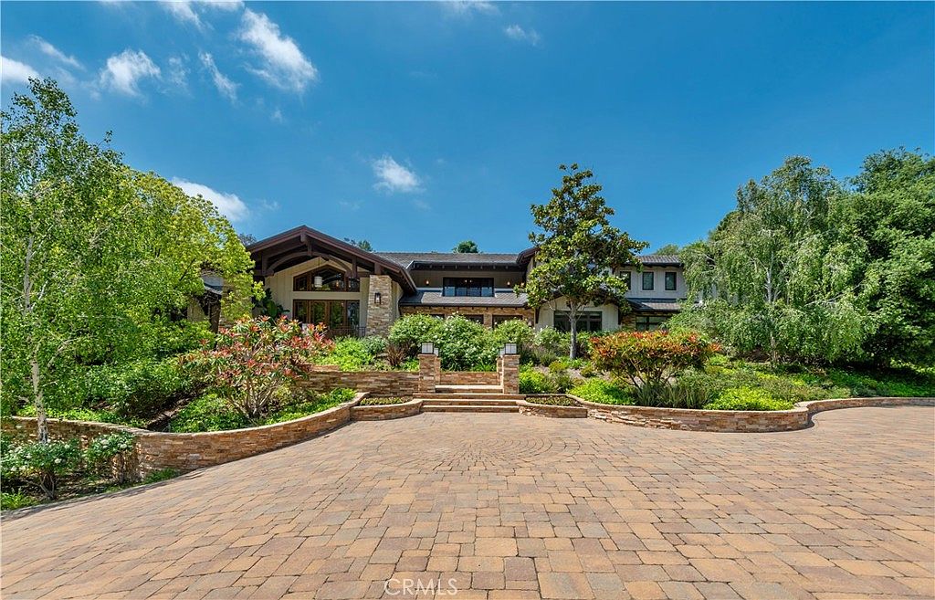 This grand, multi-level estate features a sophisticated architectural design with a blend of stone and wood elements, set against a backdrop of lush, mature trees. A wide, paved circular driveway leads up to a tiered stone staircase that serves as an inviting entryway to the home. The property exudes a sense of luxury and privacy, enhanced by the meticulously landscaped terraced gardens and the bright, clear blue sky.