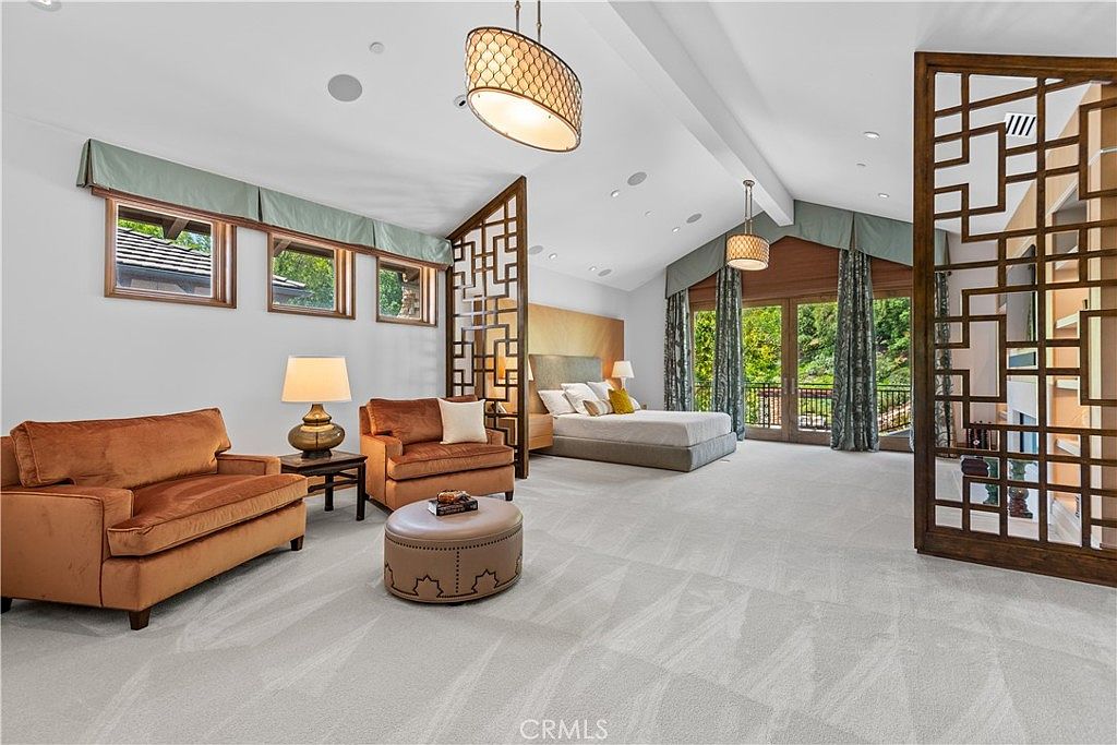 This expansive primary bedroom features a sophisticated open-concept layout with vaulted ceilings and elegant wooden room dividers that create distinct living and sleeping zones. The room is furnished with a plush velvet sofa set, a central ottoman, and a large bed, all set against a neutral color palette with warm wood accents. Large windows and glass doors allow for abundant natural light, offering a serene and luxurious atmosphere.