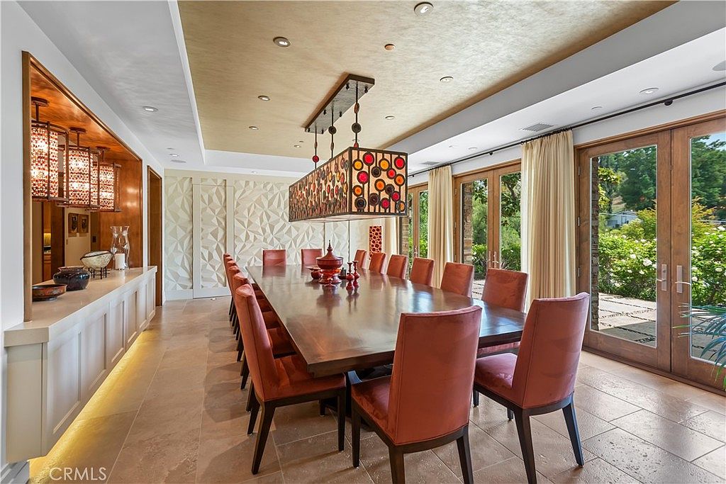 This elegant dining room features a long, polished wooden table surrounded by twelve upholstered chairs, perfect for formal gatherings. A striking, multi-colored circular pendant light fixture hangs above the table, while a textured, geometric accent wall adds depth and sophistication to the space. Large glass doors provide ample natural light and a seamless view of the lush greenery outside, creating an inviting and luxurious atmosphere.