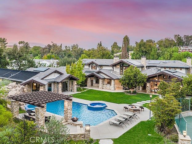 This stunning aerial perspective captures the expansive rear exterior of a luxury estate at sunset, showcasing a sprawling multi-winged residence with stone accents and a dark-tiled roof. The property features a large, sparkling swimming pool with an integrated spa, a shaded pergola lounge area, and a fire pit surrounded by comfortable seating. Lush green lawns and mature landscaping frame the home, creating a serene and sophisticated outdoor living environment.
