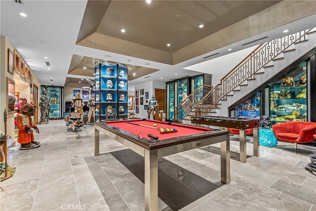 This expansive, custom-designed basement serves as a dedicated entertainment and memorabilia gallery, featuring a prominent pool table as the central focal point. The room is adorned with extensive glass display cases housing a vast collection of sci-fi figures and props, complemented by a grand staircase with decorative metal railings. The space is finished with neutral stone flooring and recessed lighting, creating a sophisticated yet playful atmosphere for collectors and guests.