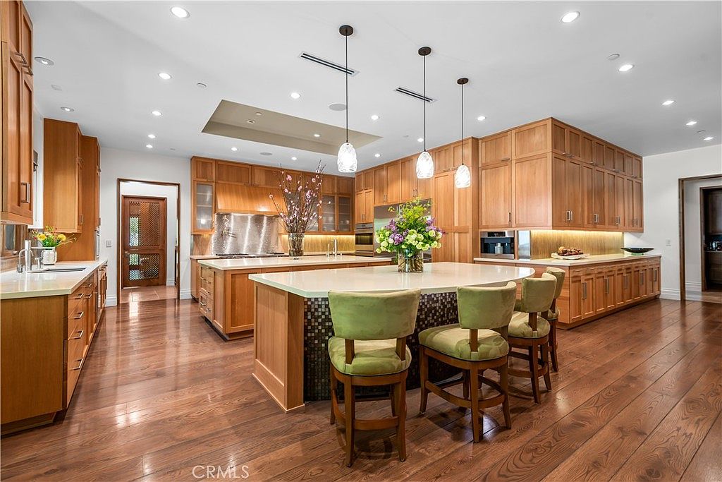 This expansive, high-end kitchen features warm wood cabinetry, a large central island with seating, and sleek light-colored countertops. The space is illuminated by elegant pendant lighting and recessed ceiling lights, creating a bright and inviting atmosphere. The perspective is a wide-angle shot that highlights the open layout, premium finishes, and the seamless flow between the cooking and dining areas.