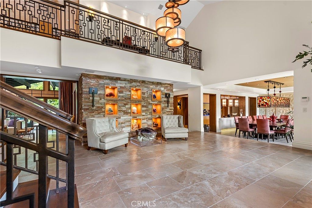 This grand, open-concept living area features a dramatic double-height ceiling and a striking stone accent wall with built-in illuminated display niches. The space is anchored by a pair of elegant, light-colored armchairs and a polished stone coffee table, all set upon large-format travertine tile flooring. A sophisticated wrought-iron staircase railing overlooks the room from the mezzanine, while a formal dining area is visible in the background, creating a luxurious and airy atmosphere.
