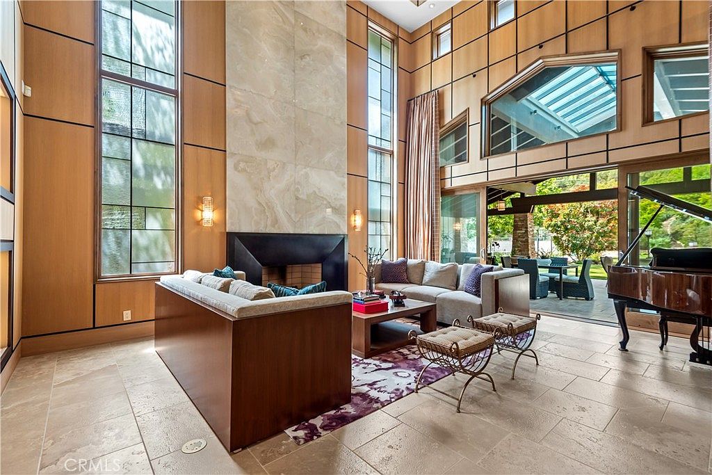 This expansive, double-height living room features a dramatic floor-to-ceiling stone fireplace as its centerpiece, framed by elegant wood-paneled walls and towering windows. The space is furnished with a plush, modern sofa arrangement and a grand piano, creating a sophisticated atmosphere that seamlessly transitions to an outdoor patio through large glass doors. The combination of natural light, high-end materials, and open-concept design conveys a sense of luxury and architectural grandeur.