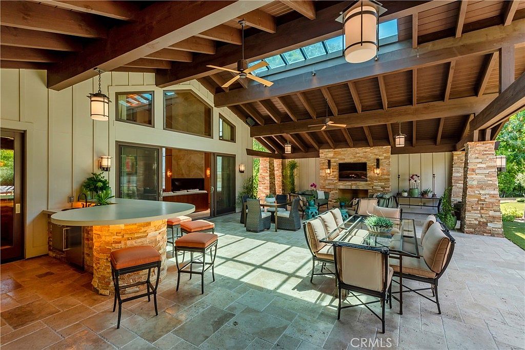 This expansive, covered outdoor patio features a rustic-modern design with exposed wooden beams, a stone fireplace, and a built-in bar area. The space is furnished with a formal dining set and additional lounge seating, all set against a backdrop of natural stone and large glass sliding doors that connect to the interior. The open-air layout, complete with a skylight and ceiling fans, creates a luxurious and inviting atmosphere perfect for year-round entertaining.