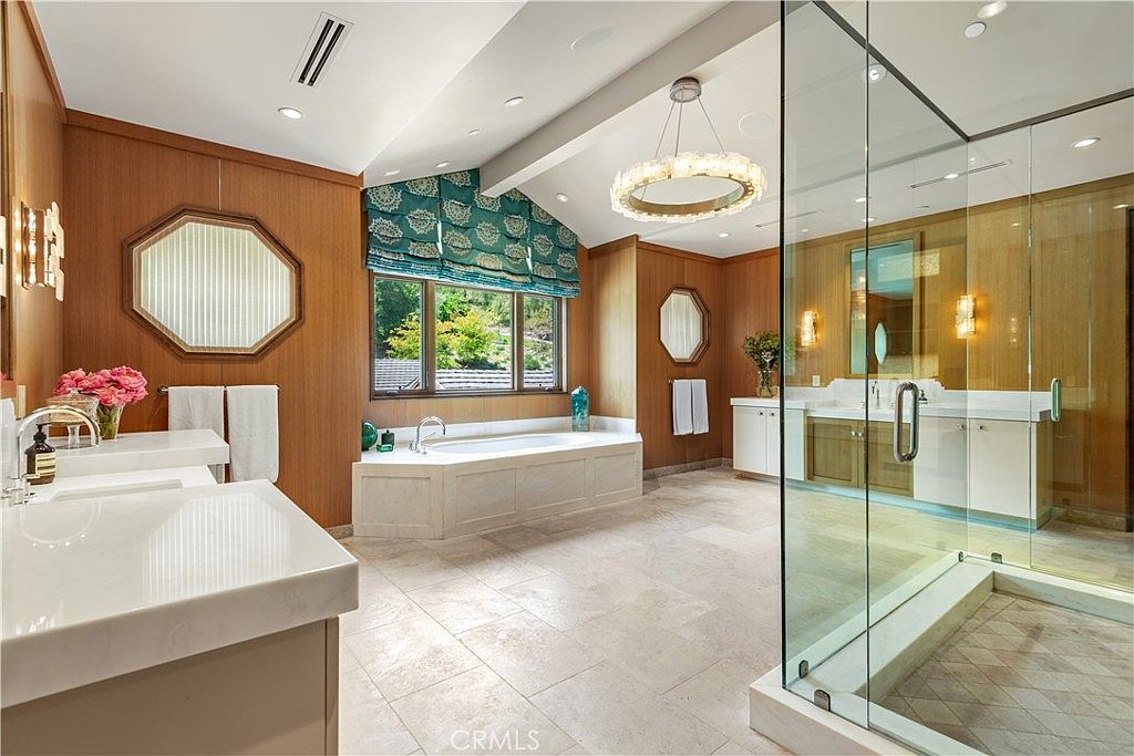 This luxurious primary bathroom features warm wood-paneled walls, a deep soaking tub positioned beneath a window with patterned valances, and a spacious glass-enclosed walk-in shower. The room is illuminated by a sophisticated tiered chandelier and recessed lighting, highlighting the elegant stone flooring and dual vanity setup. The overall aesthetic is one of refined, classic comfort with a bright and airy atmosphere.