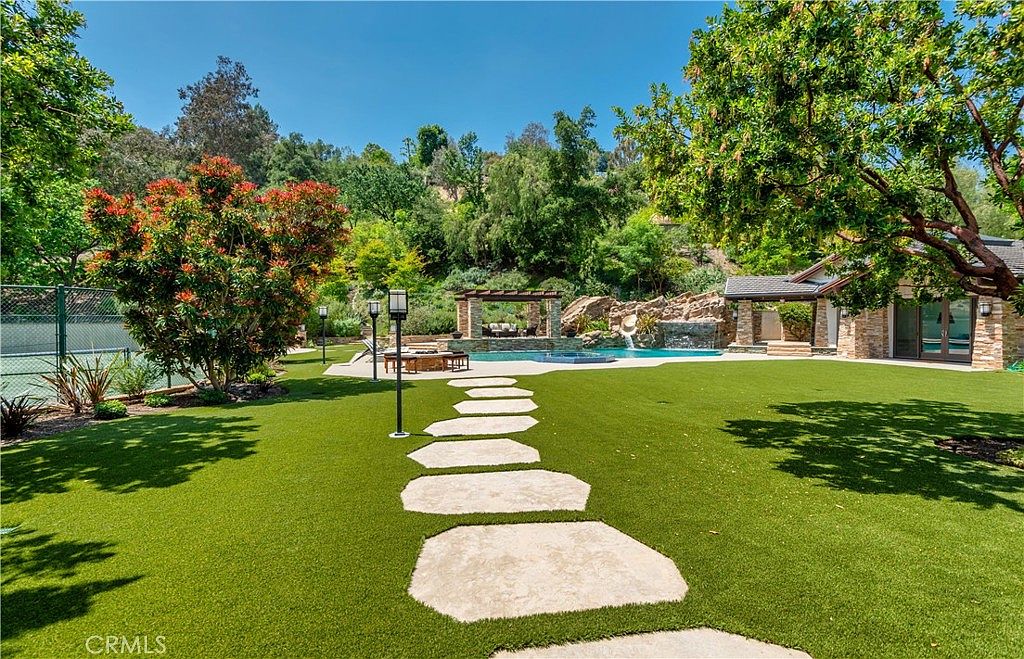 This expansive backyard features a lush green lawn with a stone paver pathway leading toward a resort-style swimming pool and spa area. The scene is framed by mature trees and a hillside backdrop, with a pergola providing a shaded lounge space near the water. The overall impression is one of luxury, privacy, and a perfect setting for outdoor entertaining.
