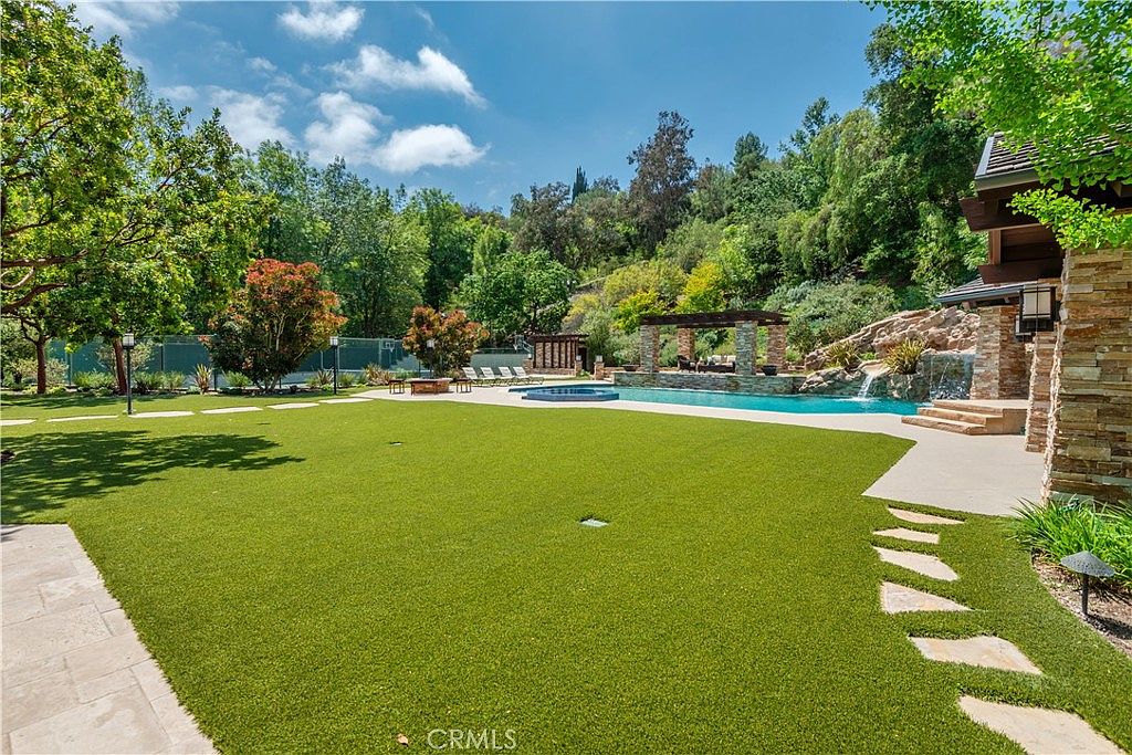 This expansive backyard features a pristine swimming pool with a natural rock waterfall feature and an adjacent spa. A large, manicured artificial turf lawn provides a clean, open space for recreation, while a covered pergola offers a shaded lounge area. The property is surrounded by lush, mature trees and hillside landscaping, creating a private and serene resort-like atmosphere.