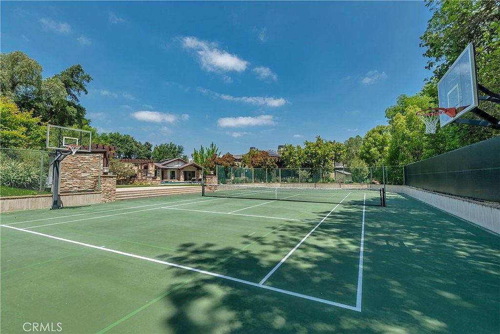This expansive outdoor space features a multi-purpose sports court with both tennis and basketball facilities, set against a backdrop of lush green trees and a clear blue sky. The court is well-maintained with crisp white lines, and a stone-accented wall adds a touch of rustic elegance to the perimeter. The perspective is wide and inviting, showcasing a premium recreational area perfect for active living.