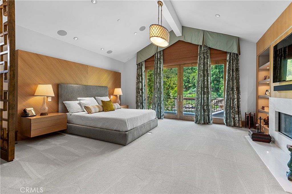 This spacious primary bedroom features a contemporary design with a large, upholstered bed centered against a warm wood-paneled accent wall. The room is highlighted by a vaulted ceiling, a sophisticated chandelier, and floor-to-ceiling glass doors that open to a private balcony with lush greenery views. A built-in fireplace and shelving unit add a cozy, luxurious touch to the expansive, carpeted living space.