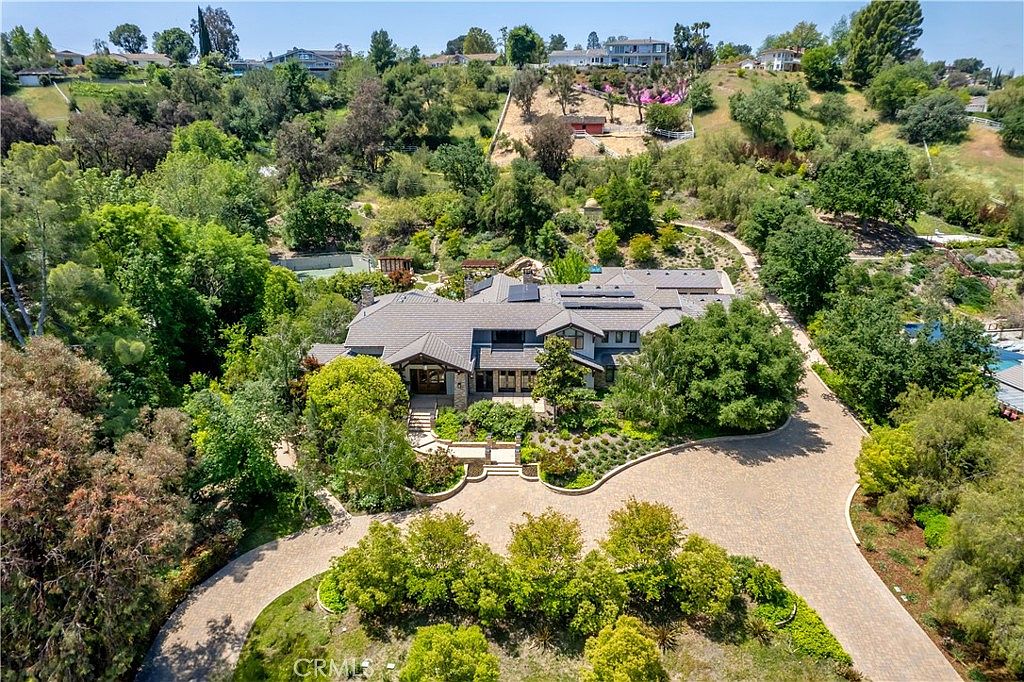 This high-angle aerial view captures a sprawling, luxurious estate nestled within a lush, hilly landscape. The property features a large, multi-gabled residence with a paved circular driveway, surrounded by mature trees, manicured gardens, and expansive greenery. The perspective highlights the home's impressive scale and its secluded, park-like setting, emphasizing both privacy and architectural elegance.