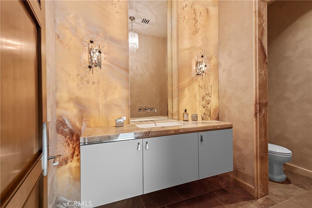 This elegant powder room features a striking vanity with a polished stone countertop and a sleek, light-colored cabinet. The walls are adorned with luxurious, warm-toned marble slabs, complemented by sophisticated starburst-style wall sconces and a large, frameless mirror. The space is finished with a modern toilet and dark tile flooring, creating a refined and high-end atmosphere.
