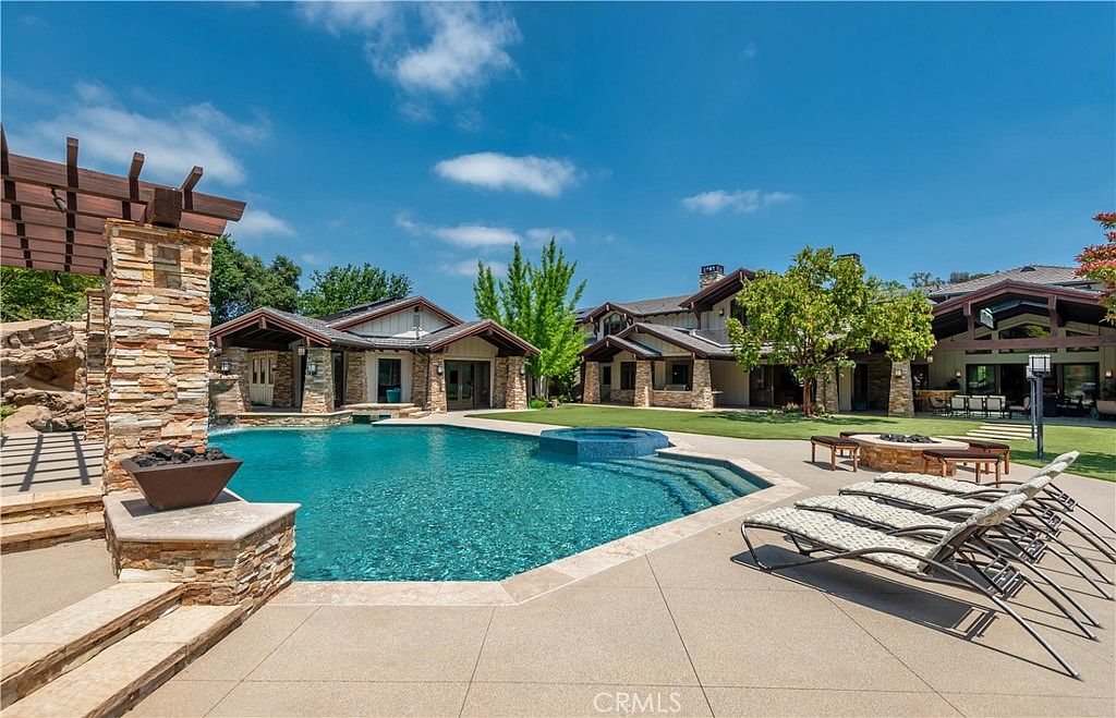 This expansive backyard features a large, custom-shaped swimming pool with an integrated spa and stone-tiled steps. The surrounding patio is furnished with several lounge chairs and a stone fire pit, all set against the backdrop of a sprawling, multi-gabled luxury residence with stone accents. The scene is captured from a low-angle perspective, emphasizing the resort-like atmosphere and the seamless transition between the indoor living spaces and the outdoor entertainment area.